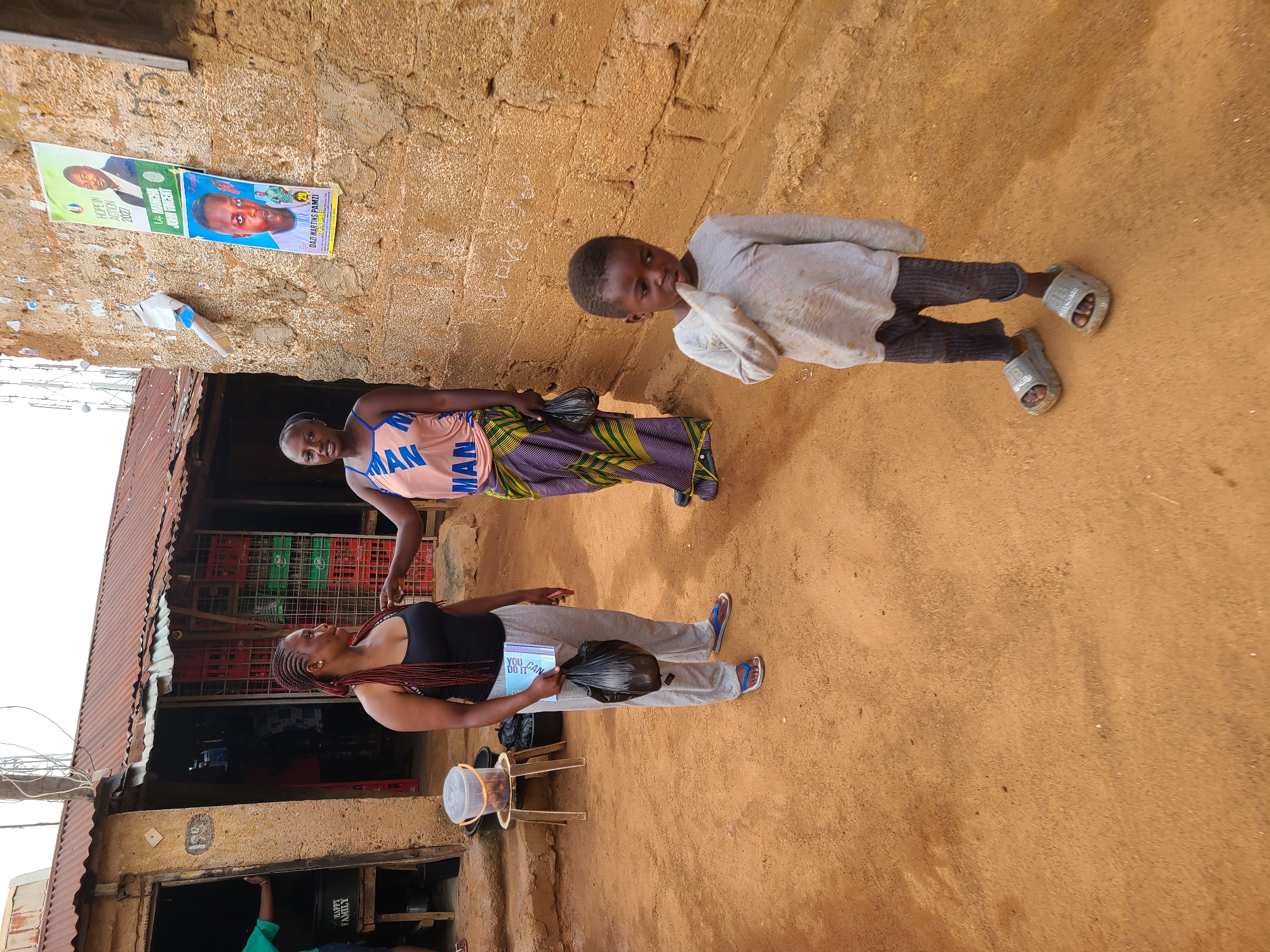 This image, captured during the daytime in Jos, Nigeria, depicts an outdoor scene featuring three individuals against a backdrop of a building and a light brown wall. The ground is reddish-brown earth.
To the left, a structure with a corrugated metal roof suggests a small shop or dwelling, its interior revealing stacks of red and green plastic crates. In the foreground, a woman with braided hair, wearing a black tank top, grey sweatpants, and blue and white sandals, stands holding a book titled "YOU CAN DO IT" and a black plastic bag. She looks to the left, away from the camera. A white plastic bucket rests on a wooden stand near her.
Beside her, a second woman, dressed in a pink top featuring the repeated word "MAN" and a patterned skirt, holds a black plastic bag and looks towards the camera. To the right, a young boy in a light-colored long-sleeved shirt, dark pants, and grey sandals stands, with a finger near his mouth, looking to the left.
The wall behind the individuals features two political campaign posters. One poster displays a candidate named "U MANCHA JOHN VINCENT" and the text "HOPE IN ACTION 2027". The other poster shows "DAZI MARTINS PAMZI" with the number "29". Below the shop's entrance, the words "HAPPY FAMILY" are visible. The scene captures a moment of daily life in the community under clear, bright skies.
