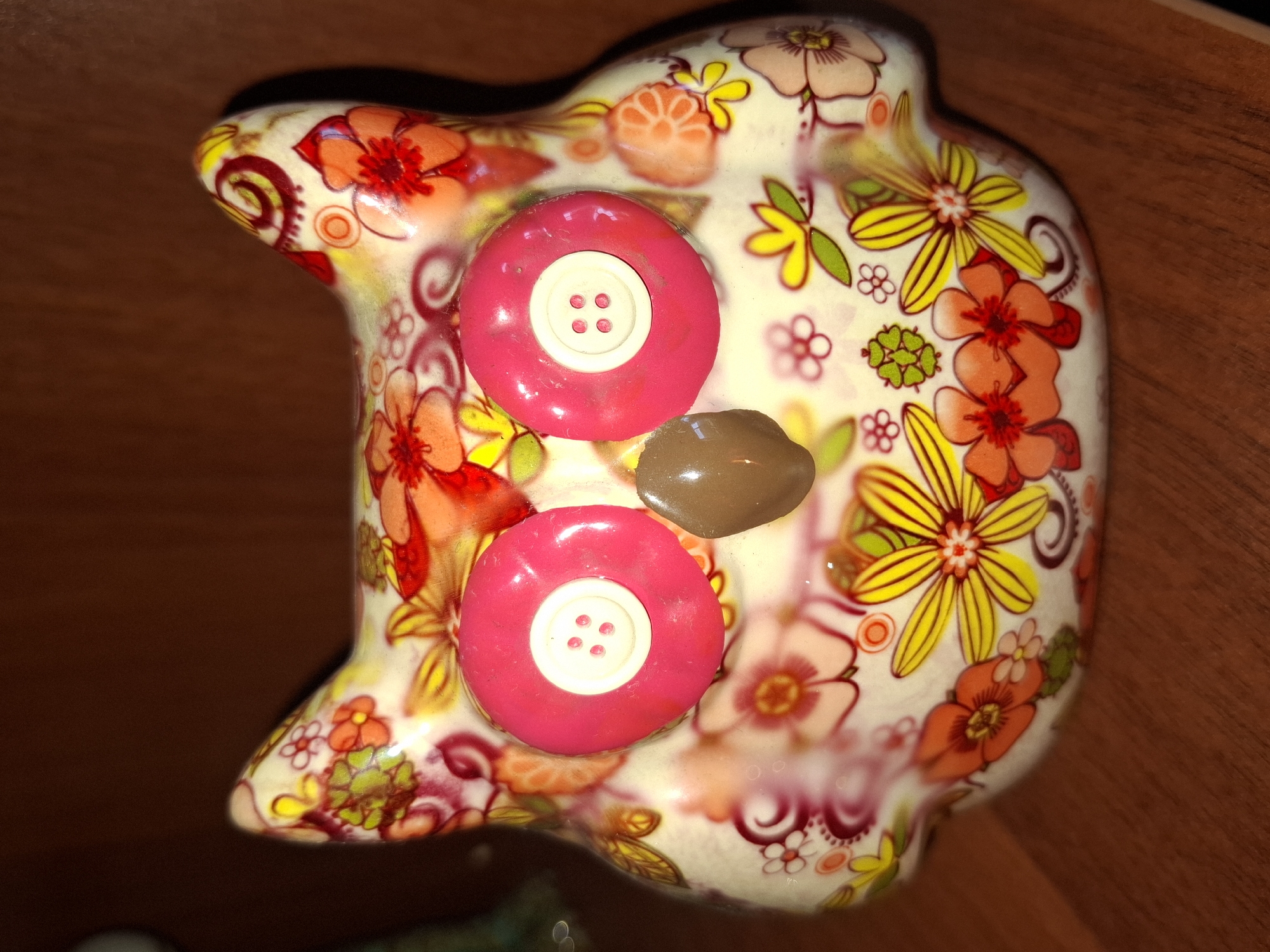 A close-up, eye-level, slightly downward-angled shot shows a ceramic owl figurine. The figurine is decorated with a vibrant floral pattern in shades of pink, orange, yellow, and green on a white background. It has two large, round, bright pink eyes with white button-like centers. A small, oval, brown object, resembling a bean, is placed in the center, between the eyes, forming the beak. The figurine is situated on a wooden surface, likely a shelf or table. The background is a dark, blurred brown. The lighting creates some glare on the glossy surface of the figurine. The image does not contain any contextual information about its location in Almaty, Kazakhstan.