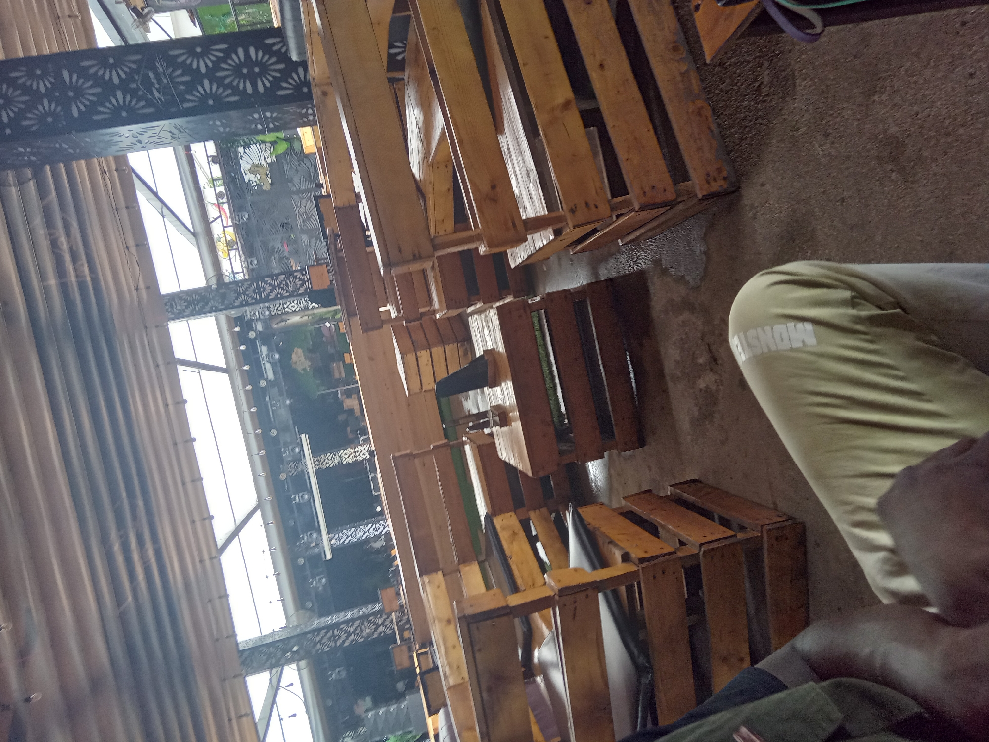 The image depicts a semi-enclosed outdoor or open-air casual dining establishment or lounge, located in Howayi, Nigeria. The area features numerous seating arrangements constructed from light-colored, natural wood pallets, serving as both tables and benches. Many of the benches are fitted with dark grey or black cushions. The floor is a concrete or tiled surface, exhibiting multiple wet patches. Overhead, a roof structure composed of corrugated panels incorporates several skylights, allowing natural light to enter. Supporting the roof are dark-colored structural pillars adorned with intricate, floral-patterned cutouts. In the background, similar patterned screens or walls are visible, along with mounted lighting fixtures and string lights strung across the ceiling. In the immediate foreground, a person is seated, partially visible from the chest down. They are wearing a dark shirt under a green or khaki vest and light-colored trousers with the word "MONSTER" printed vertically on the left leg. Their arm is resting on their lap. On a central table, a dark stand, possibly for menus or condiments, is present. The overall scene appears to be devoid of active patrons beyond the partially visible individual in the foreground.