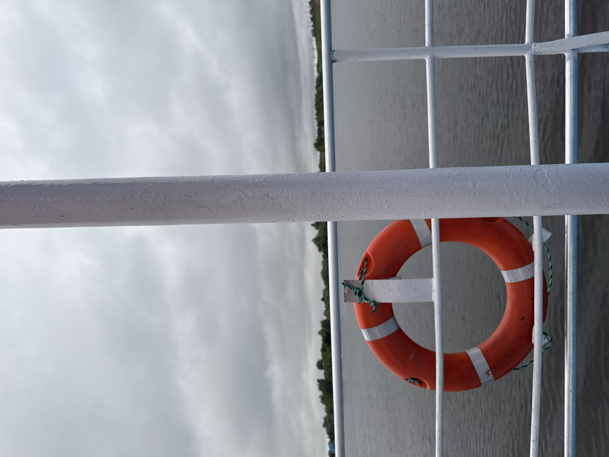The image depicts a close-up view of a life preserver attached to a white railing on a boat. The life preserver is bright orange with two reflective white bands. A green rope is tied to the life preserver and secured to a white pole. The railing is made of horizontal white bars. In the background, a body of water extends to the horizon, where a treeline is visible under a cloudy, overcast sky. The scene suggests a maritime environment, likely on a river or lake, given the calm water and visible treeline. The location of Шебунино, Russia, is provided, implying this scene is situated within or near this area, which is known for its natural landscapes and waterways. There are no people or animals visible in the image.