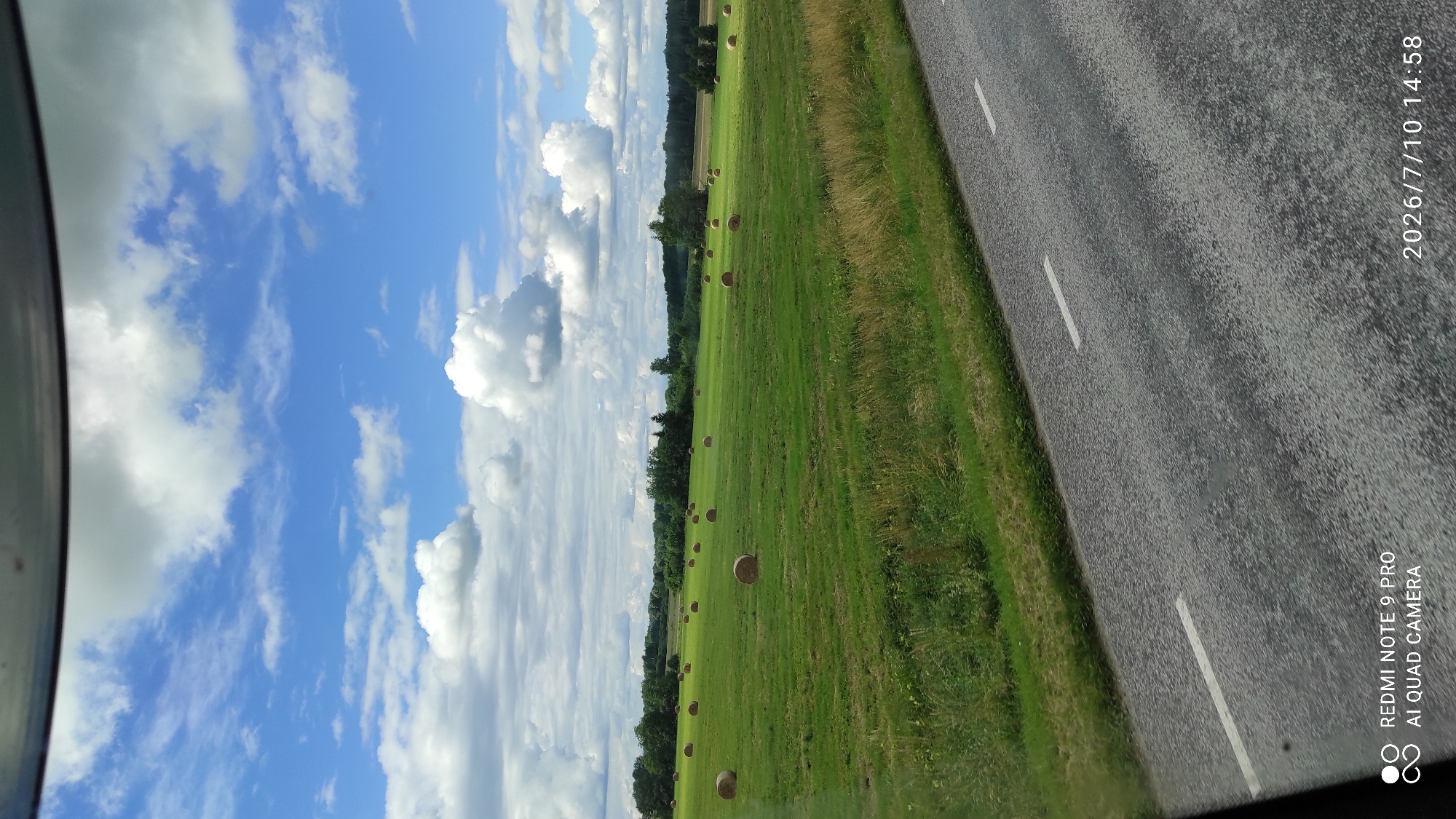 The image captures a pastoral scene in the vicinity of Olustvere, Estonia, on July 10, 2026, at 14:58. A vast, green field stretches across the midground, dotted with numerous round hay bales. The field is bordered by a dense line of trees in the background, with a subtle hint of cultivated land or further fields visible beyond the treeline. The sky above is a vibrant blue, adorned with a scattered arrangement of white, cumulus clouds, suggesting fair weather. In the foreground, a paved road with white lane markings is visible, implying the vantage point is from a vehicle traveling along this route. The overall impression is one of rural tranquility and agricultural activity under a summer sky.
