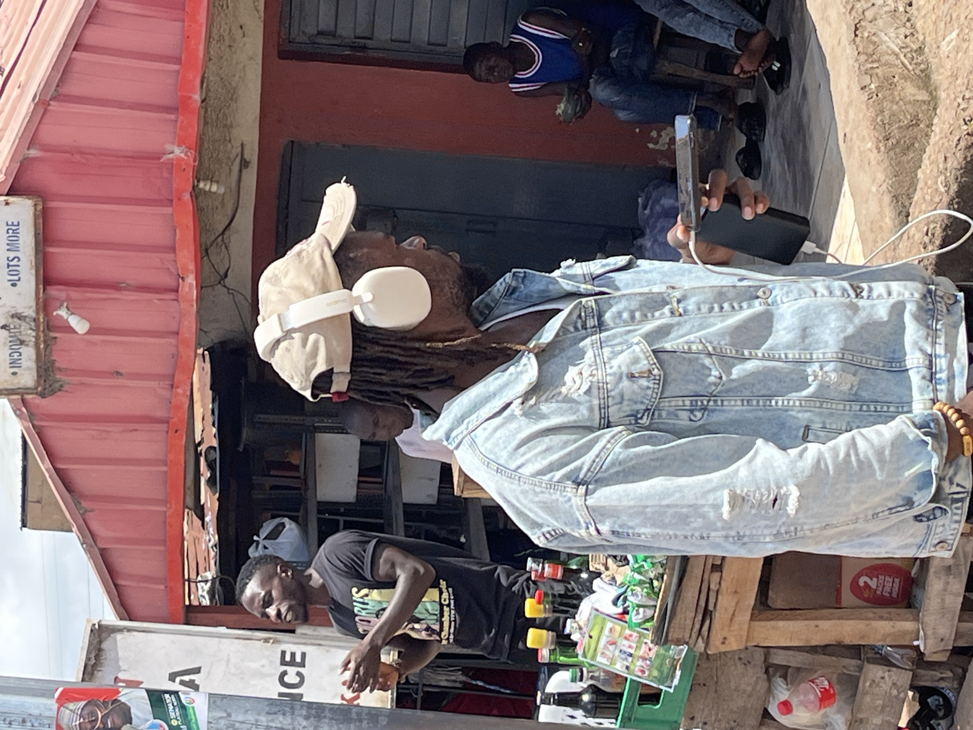 A medium shot captures a bustling street scene in Gura Top, Nigeria. The foreground is dominated by a man wearing a light blue distressed denim jacket and a beige baseball cap with white headphones. He holds a smartphone in his right hand, connected by a white cable to a black power bank in his left. His gaze is directed to the right, out of frame. Behind him, a stall displays various bottles and packaged goods. To his left, another man in a black t-shirt with text and graphics is visible, holding a glass of amber liquid and gesturing with his hands. In the background, a man in a blue tank top with white trim sits holding a piece of fruit. The environment appears to be a street market with red corrugated metal roofing visible above.
