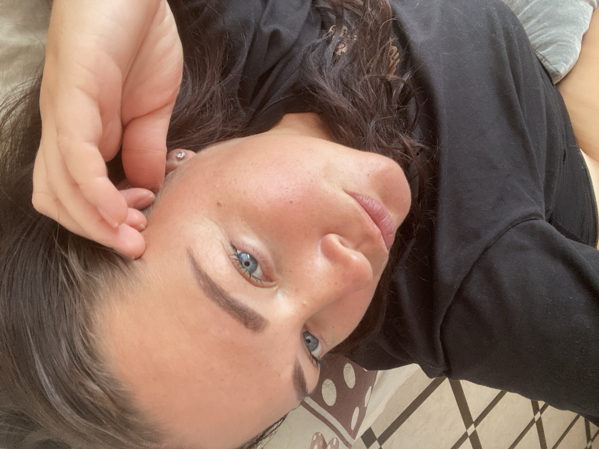 The image shows a close-up, vertically oriented, outdoor selfie of a woman lying down. She is wearing a black long-sleeved shirt and has long, dark, wavy hair. Her face is turned upwards, and her blue eyes are looking towards the camera. Her left hand is touching her face near her temple. The background shows a patterned pillow and a tiled floor, suggesting an indoor setting. The lighting is natural and appears to be daylight. The woman has fair skin with some freckles and is wearing subtle makeup. There is no discernible location information that could pinpoint it to Sochi, Russia.