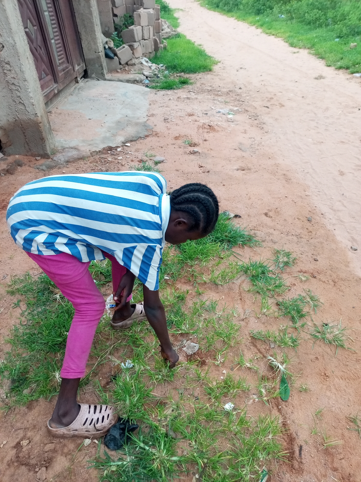 An individual, likely a child, is bending down to pick something off the ground in Tempure, Nigeria. They are wearing a blue and white striped shirt and pink pants, with light-colored sandals on their feet. The ground is a mixture of sandy soil and sparse green grass, with some scattered debris. In the background, there is a concrete structure with a wooden door, some stacked bricks, and a dirt path flanked by green vegetation. The overall environment appears to be a rural or semi-urban outdoor setting.