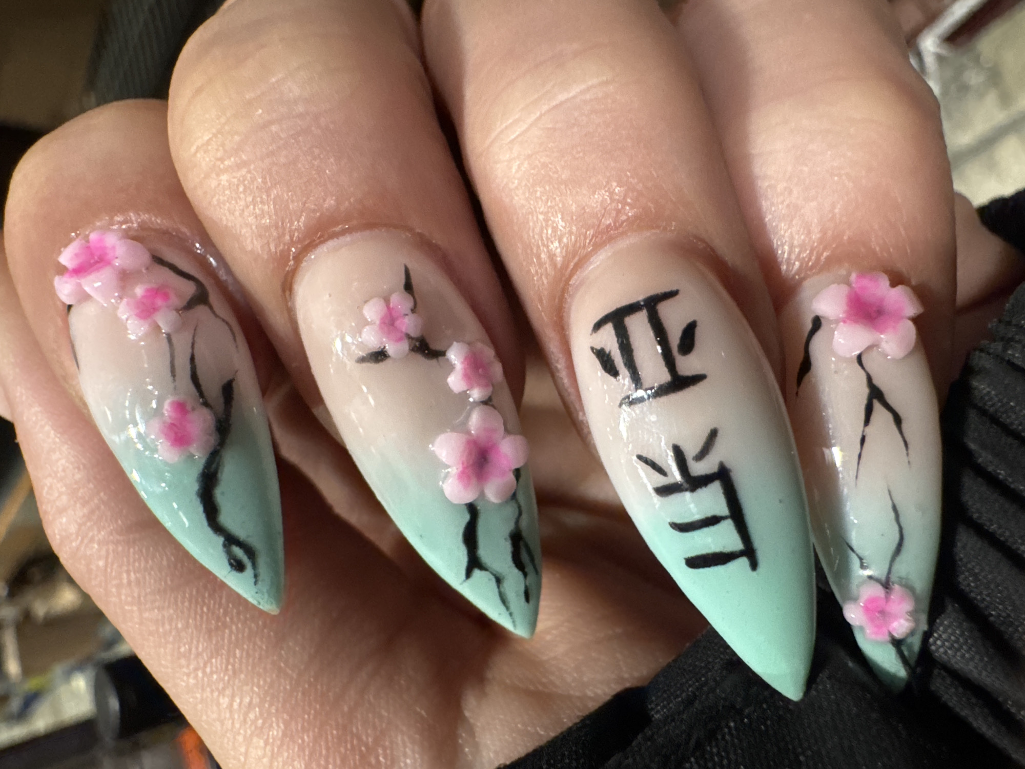 Close-up image of a hand displaying four fingernails with detailed designs. The nails are long, pointed, and have a gradual color gradient from a milky white at the base to a mint green at the tips. Three nails have a cherry blossom design with black stems and small pink flowers; the flowers on the first and fourth nails are three-dimensional. The middle nail features two black, stylized Chinese characters. The background shows a blurred black garment and a dark, out-of-focus surface. The location is unknown.