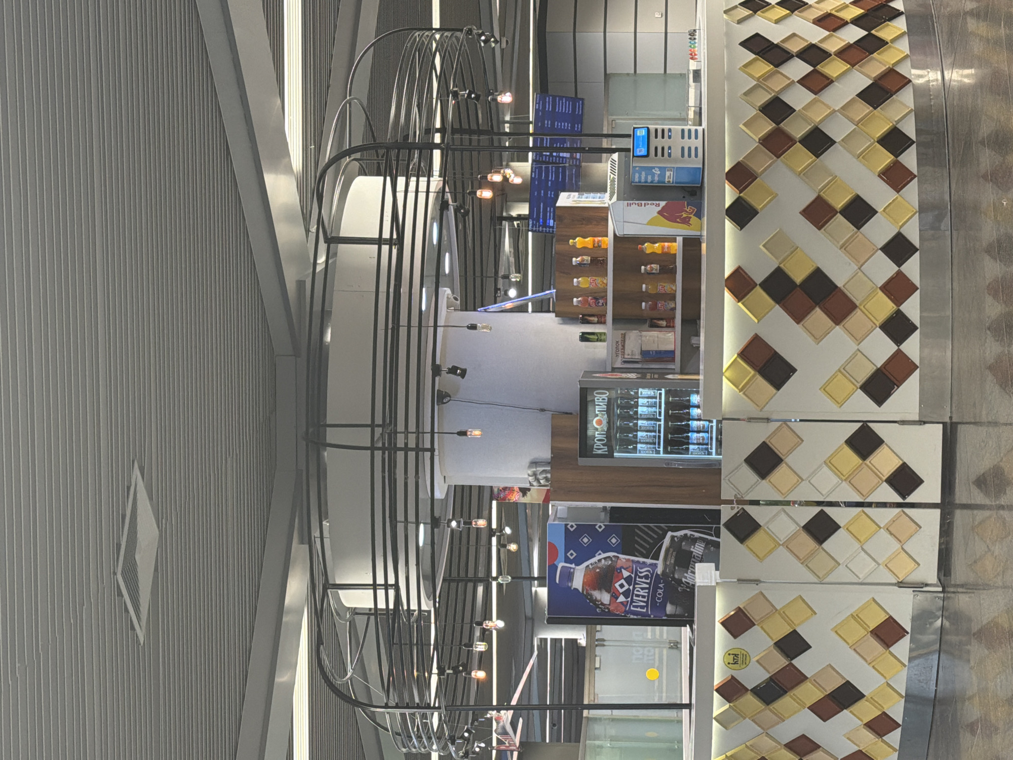 The image shows an interior scene, likely a concession stand or small shop, possibly in an airport or train station. A curved counter with a decorative tiled facade is prominent in the foreground. Behind the counter, various items are displayed on shelves and in a refrigerated display case, including bottles of drinks and a Red Bull cooler. The background features a large pillar, a circular metal structure with hanging lights, and a ceiling with a slatted design and a ventilation grille. A digital display board is visible in the upper right portion of the frame. There are no people in the image.