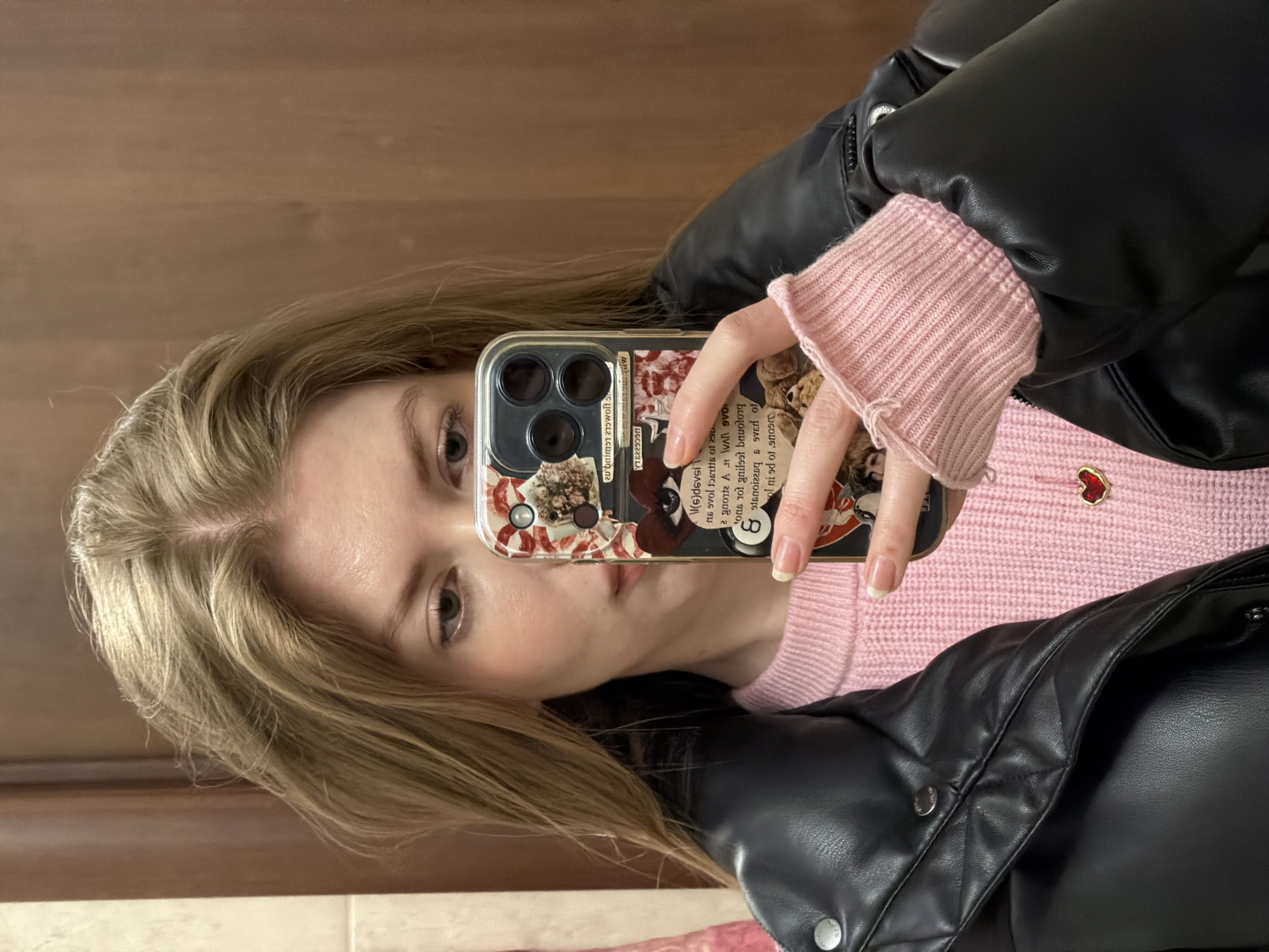 A young individual is captured in a close-up selfie, holding a smartphone. The subject's head is tilted, and they look directly towards the camera with light-colored eyes and a neutral expression. Their hair is long, light brown or blonde, and appears to be parted on the side.
They are wearing a light pink ribbed knit sweater, visible at the neckline and cuffs. A small, red heart-shaped pin with a gold border adorns the left side of the sweater. Over the sweater, a black puffer jacket, possibly made of leather or faux leather, is worn, featuring visible zippers and snap buttons. The individual's fingers display a French manicure with light tips.
The smartphone is encased in a clear cover decorated with a collage of various elements, including what looks like dried flowers or botanical fragments, patterned paper, and snippets of text. A distinct black "8 ball" design is also visible on the case. Due to the selfie being a reflection or taken with a front-facing camera, the text on the phone case appears reversed. Visible reversed text fragments include "20brime", "VT6229590", "(6)devsi", "16 of 290", "agrotte At VAI 970", "Lons tot gail99 bravotonq", "ol ni ed of anoere", and "ot".
The background consists of a plain wooden surface, likely a wall or door, and a small strip of light-colored tiled floor at the bottom of the frame, indicating an indoor setting. The lighting is even.