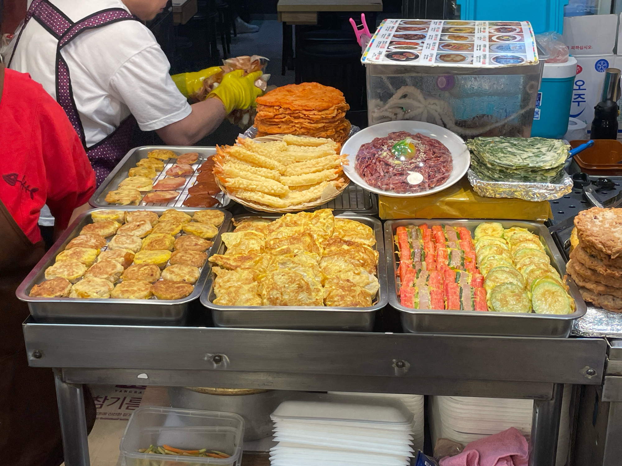 This image captures a street food vendor stall in Seoul, South Korea, likely in a busy marketplace or commercial district. The stall is laden with various Korean street food items, neatly arranged in metal trays. In the foreground, a person wearing yellow gloves is visible, attending to the food, suggesting an active food preparation or serving area.
The food displays include an assortment of fried items: a tray of round, golden-brown fritters; a tray of thinly sliced, orange-colored food; a basket of crispy fried shrimp; a platter of what appears to be steak tartare with a quail egg on top, covered in plastic wrap; and a tray with green vegetable pancakes or jeon. Another tray contains colorful rolls made of red crab sticks and green vegetables, alongside sliced green squash pancakes. To the right, a stack of large, round, browned patties is piled high.
Below the main food display, white styrofoam containers are stacked, ready for customers. A transparent plastic container at the bottom left holds some chopped vegetables, possibly for garnishing or side dishes. The background shows elements of a typical urban environment with tables and chairs, suggesting a dining area or a restaurant setting. The overall impression is one of a bustling food stall, showcasing a diverse range of popular Korean street food.