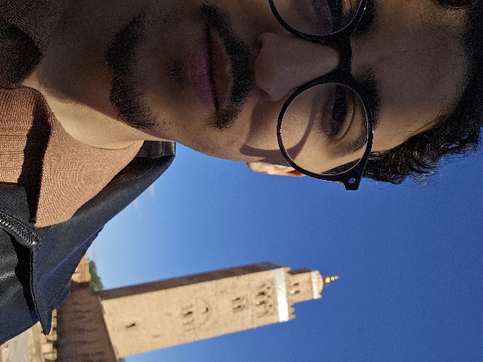 The image displays a close-up selfie of a person, positioned at an angle, with a prominent architectural landmark and clear blue sky in the background. The original image is rotated 90 degrees counter-clockwise.
The subject is a person whose face is partially visible, from just below the mouth to the top of the head. They have dark, curly hair and dark, well-defined eyebrows. They are wearing round, black-rimmed glasses, and their right eye (on the left side of the frame from the viewer's perspective when oriented upright) is clearly visible through the lens. A dark mustache and goatee are present. The person's lips are slightly parted. They are dressed in a light brown or terracotta-colored collared top, layered with a black jacket, possibly leather, which has a visible zipper on the upper chest area. Strong sunlight casts distinct shadows on the person's face, particularly under the chin and around the facial hair, suggesting bright daytime conditions.
In the background, under a vast, cloudless blue sky, stands a tall, reddish-brown stone or brick tower, which appears to be a minaret due to its architectural style and vertical design elements. The tower has intricate patterns or carvings visible on its facade and is topped with a golden or light-colored finial. The building is slightly out of focus, indicating a shallow depth of field, typical of a close-up photo where the background is distant.
The scene suggests a travel or tourist context, where the person is capturing a moment with a significant landmark. The clear blue sky indicates fair weather, and the strong shadows point to a sunny day, likely during late morning or early afternoon. The style of the minaret is characteristic of Islamic architecture, potentially indicating a location in a region like North Africa, possibly Marrakech, given the distinctive reddish hue of the building. No discernible text is visible in the image.