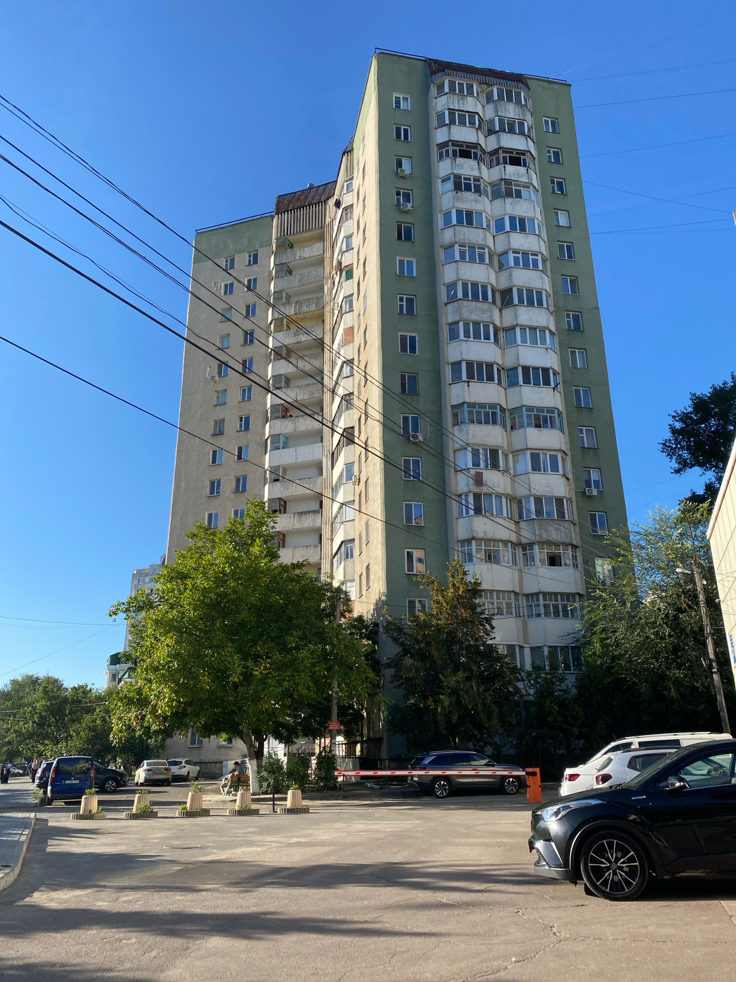 This is a street-level, eye-level, full-shot photograph taken in Chișinău, Moldova. The image features a tall, multi-story apartment building dominating the background, with its light-colored facade and green upper sections. Numerous balconies, many enclosed with windows, are visible along the building's exterior. Overhead, a network of electrical wires stretches across the clear blue sky. In the foreground and midground, a paved street or parking area is visible, lined with trees and several parked cars, including a dark-colored SUV in the immediate foreground, a white SUV behind it, and a blue minivan further down the street. A red and white barrier gate is also present. A few people are sparsely scattered along the sidewalk, one of whom is sitting on a bench under a large tree. The overall environment suggests a residential urban area.