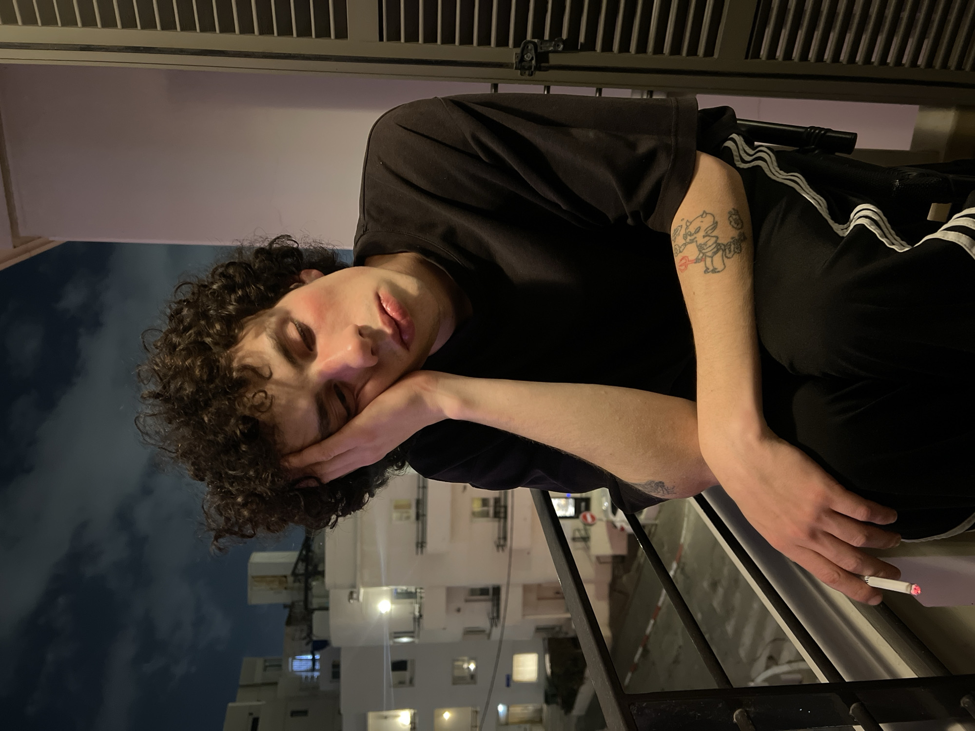 A young adult male with curly brown hair, dressed in a black t-shirt and black Adidas track pants with white stripes, is reclining with his face resting on his hand. He is holding a lit cigarette. He appears to be on a balcony in Tel Aviv, Israel, as suggested by the architecture and the buildings in the background. The time is either late in the day or at night, with streetlights illuminated in the distance. The sky is dark. The man has a tattoo on his left arm.