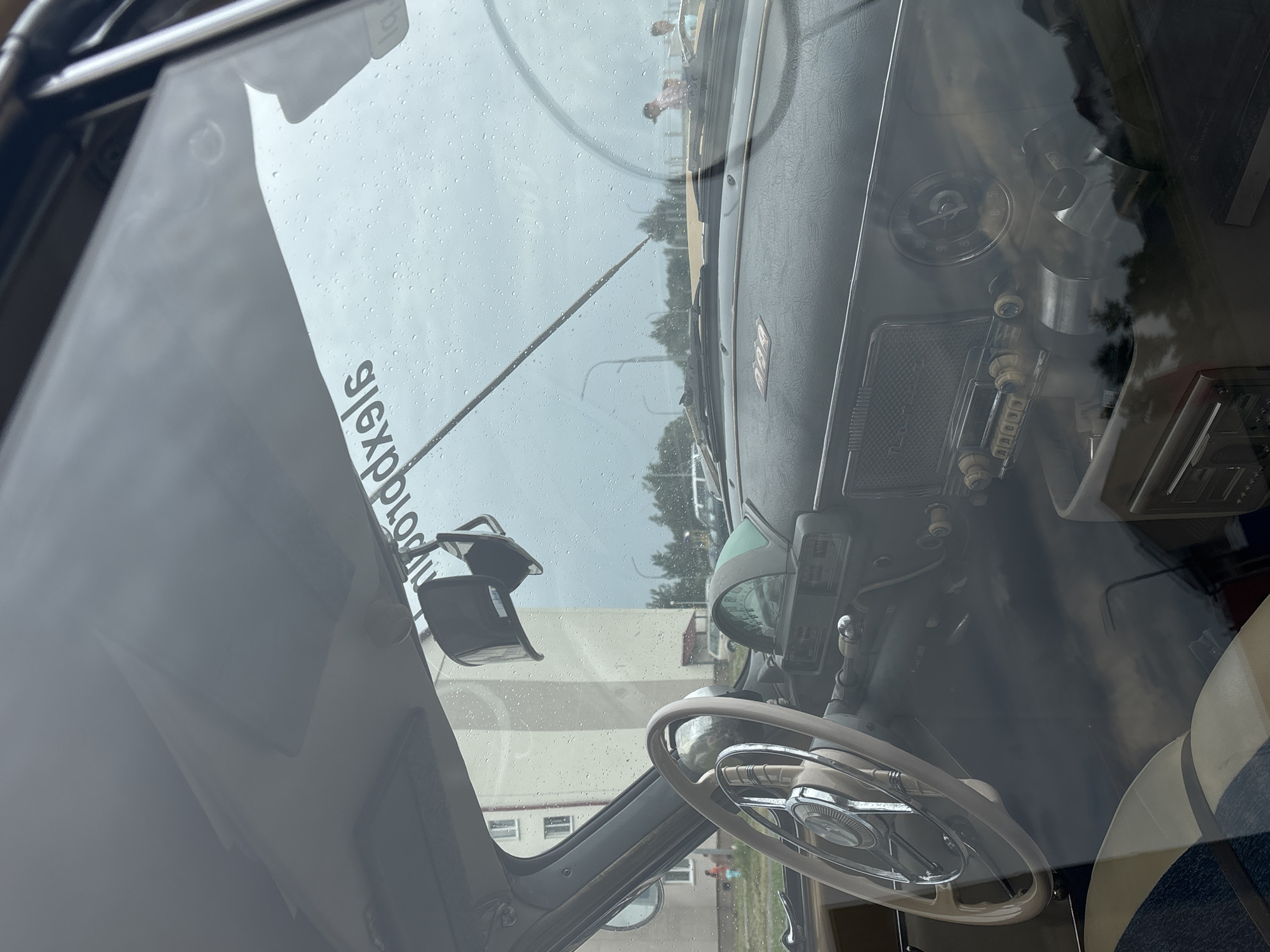An interior shot of a vintage car, likely a GAZ-21 Volga, is captured through a rain-streaked windshield. The dashboard features a prominent beige steering wheel with chrome accents and a central emblem. To the right, a cluster of vintage gauges includes a circular speedometer and a smaller dial, likely for fuel or oil. Below these, an old-fashioned radio with several knobs and buttons is visible. A hanging rearview mirror is positioned at the top of the windshield, partially obscuring a sticker with Russian text, which reads "pro ddxeig." The exterior scene visible through the windshield includes a modern apartment building with multiple windows, trees, streetlights, and other vehicles, suggesting an urban setting. Two individuals are faintly visible in the distance, one near the apartment building and another near a vehicle on the right side of the frame. The overall atmosphere is overcast and rainy. The scene is set in the city of Uzda, Belarus.