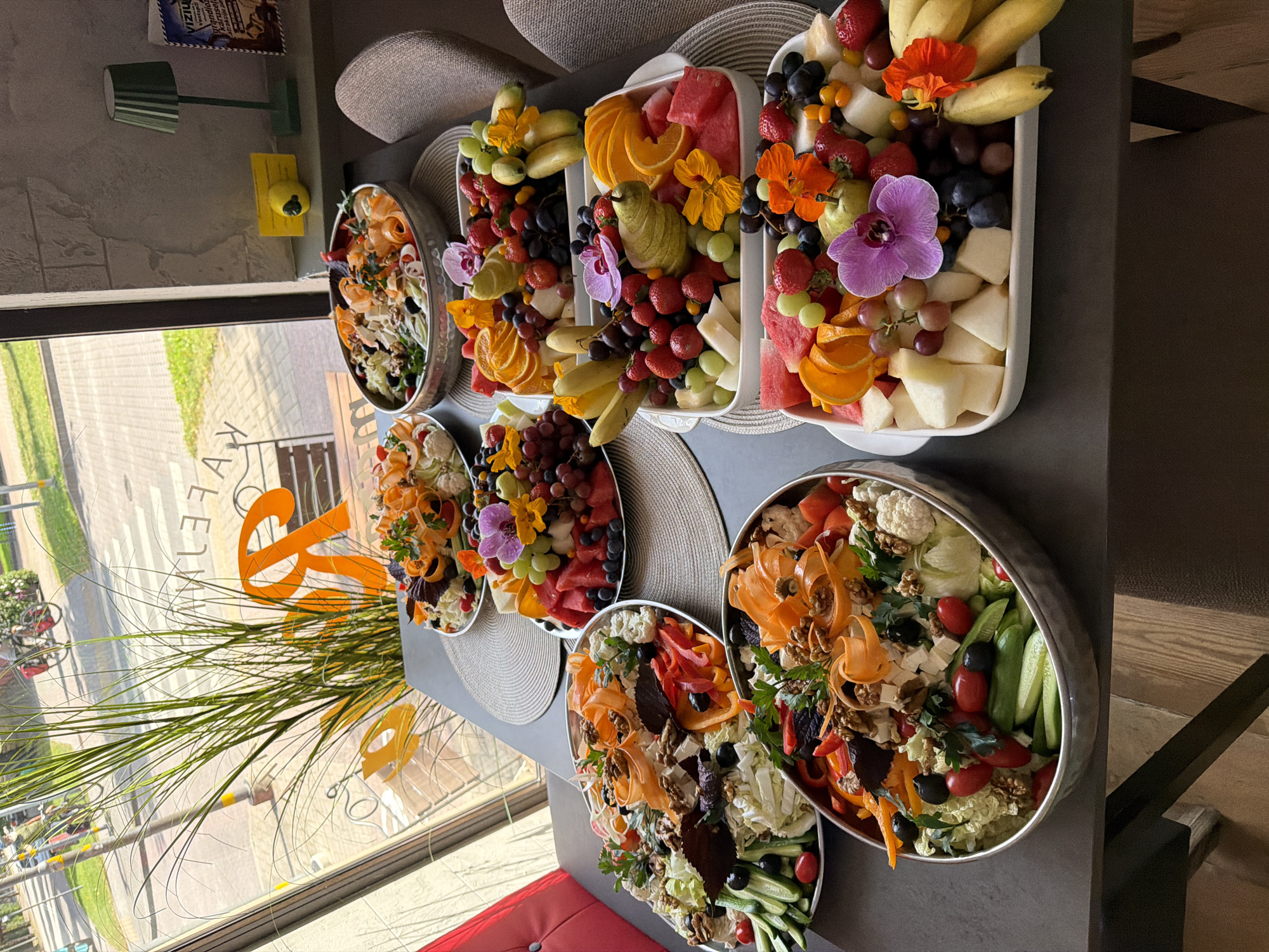 This image depicts a collection of platters filled with fresh fruits and vegetables, set on a table inside what appears to be a cafe or restaurant. The location is identified as Ventspils, Latvia, as indicated by text visible through the window, which reads "KAFA VELTA" (likely a misspelling or abbreviation of "Kafejnīca Velta" or similar).
The platters are arranged on a dark grey table with woven placemats underneath. The platters themselves vary in shape and material, with some appearing to be silver metal bowls and others white ceramic dishes.
The contents of the platters include:
* **Fruit Platters:** These are abundant with a variety of fruits such as strawberries, grapes (green and red), blueberries, watermelon, cantaloupe, oranges, bananas, and pears. Some fruit platters are also garnished with edible flowers, including purple orchids and orange nasturtiums.
* **Vegetable Platters:** At least two platters are primarily composed of vegetables. These contain elements like lettuce, cucumbers, cherry tomatoes, red bell peppers, cauliflower florets, and walnuts. Slices of carrots, thinly curled, are also prominently featured as a decorative element and ingredient.
In the background, through a large window, a street scene is visible, including a pedestrian crossing, trees, and bicycles. The lighting suggests it is daytime. Inside the establishment, a green table lamp is visible on a shelf, along with a framed item or postcard. The overall impression is that of a food service establishment preparing or presenting fresh, healthy options. No people are directly visible in the image.