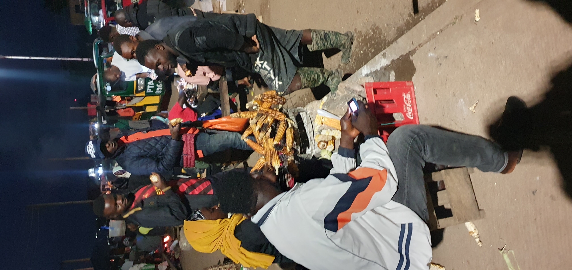 A nighttime scene in Bukuru, Nigeria, captures a street food vendor grilling corn over charcoal. Several people are gathered around, some interacting with the vendor, others observing. The foreground shows a man sitting on a stool, looking at a mobile phone. To his right, a collection of grilled corn is displayed on a metal grate over a fire. In the background, a tuk-tuk is visible, along with other market stalls and people, illuminated by streetlights and artificial lighting. The ground is paved, with a red Coca-Cola crate placed near the seated man. The atmosphere appears lively and typical of a bustling evening market.