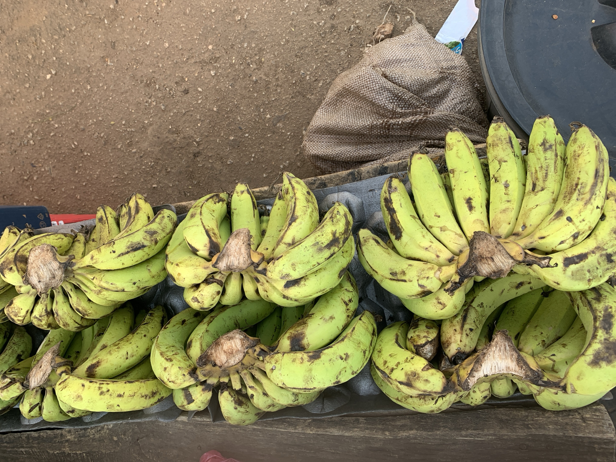 The image shows several bunches of unripe, green-yellow bananas arranged on a wooden surface. There are at least four bunches visible. Some of the bananas have brown spots. In the background, there is a brown burlap sack and a black tire. The ground is dirt. The location is New Stadium, Nigeria. There are no people visible in the frame.