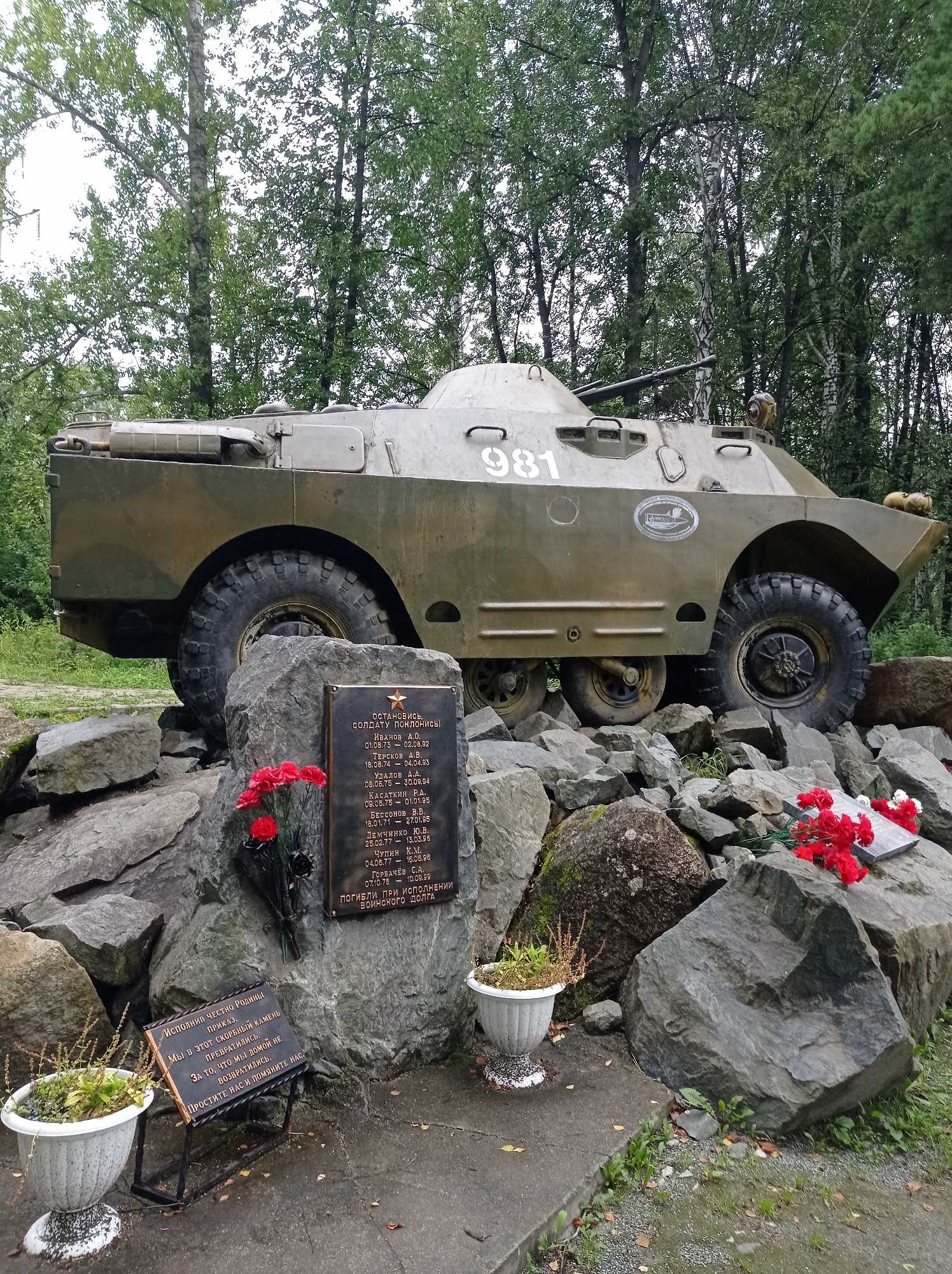 An outdoor monument features a weathered olive-drab armored personnel carrier, identified by the number "981" on its side, positioned atop a jumble of large grey rocks. In front of the vehicle, at the base of the rocks, a black plaque is affixed, bearing a list of names and dates. Red carnations are arranged in a bouquet on the plaque, with a single black flower stem also present. To the left and right of the main plaque, two white planters contain sparse green foliage. A second, smaller plaque is visible at the bottom left, mounted on a black metal stand, with a poem inscribed on it. The environment is natural, with trees and greenery forming the background, suggesting a park or wooded area. The ground consists of dirt, gravel, and some concrete paving. The overall scene appears to be a memorial site, likely dedicated to fallen soldiers, located in the city of Degtyarsk, Russia.