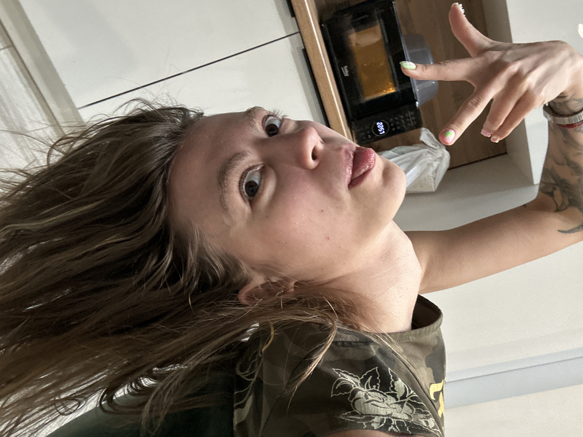 The image shows a woman with long, brown hair hanging upside down, her face tilted towards the camera. She has her tongue sticking out slightly and her eyes are open. Her right hand is raised with her fingers splayed, displaying manicured nails with green and pink designs. She is wearing a camouflage-patterned top. In the background, a microwave oven is visible on a wooden countertop. The environment appears to be a kitchen with white cabinetry. There are no visible indications of the location being in Moscow, Russia, or any specific city.