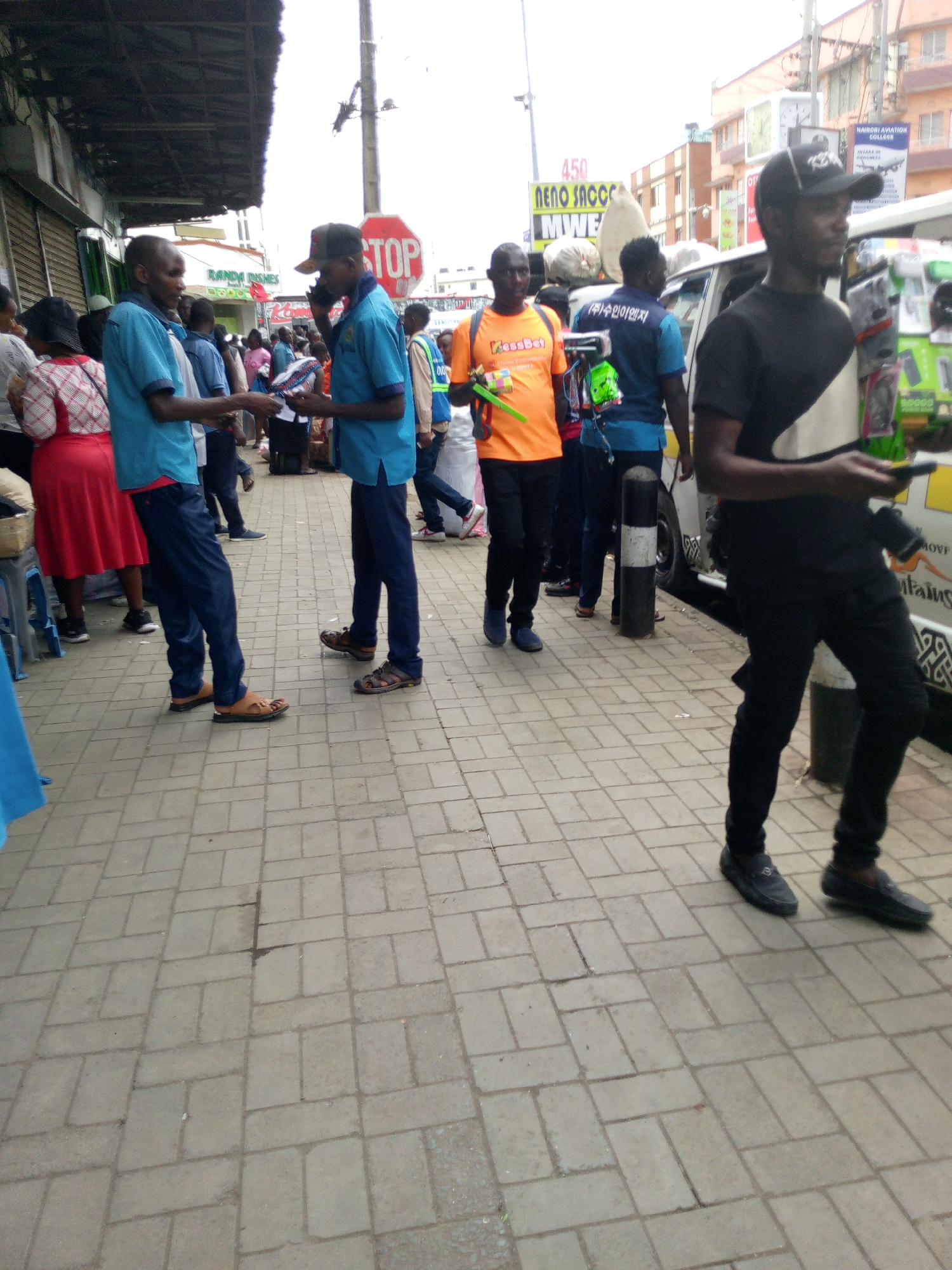 The image depicts a street scene in Nairobi, Kenya, with several individuals present on a paved sidewalk. In the foreground, a man in a black T-shirt and pants, with a baseball cap, holds objects in his hand. Further back, two men wearing light blue shirts and dark pants appear to be exchanging items. Another man is wearing an orange "MessBet" T-shirt and walking towards the left. In the background, there are signs written in English and possibly Korean. The signs are for businesses, including "NENO SACCO", "NAIROBI AVIATION COLLEGE", and "RANDA DISHES". A stop sign is also visible. The environment appears to be a bustling commercial area.