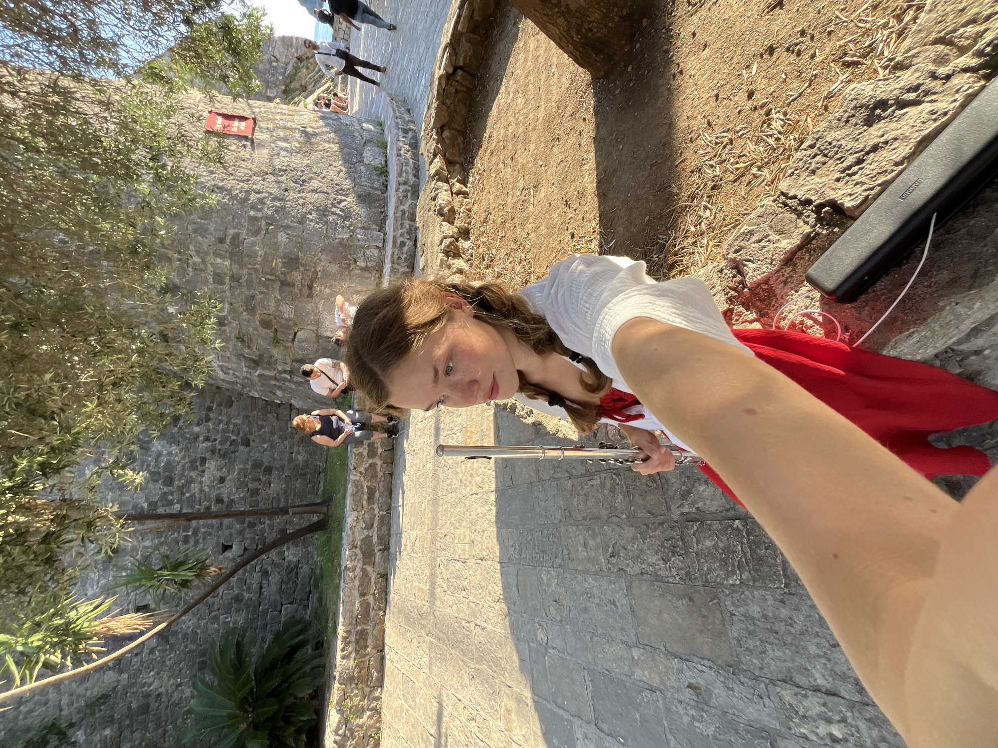 A selfie shows a young woman holding a flute while standing on a paved stone path, presumably within the city of Budva, Montenegro. The background includes an old stone wall with green trees and foliage growing along the top. In the distance, other people are walking on a pathway at the top of the wall. The woman is wearing a white shirt and a red scarf. To her right is a black rectangular flute case on the ground. The camera angle is slightly tilted.