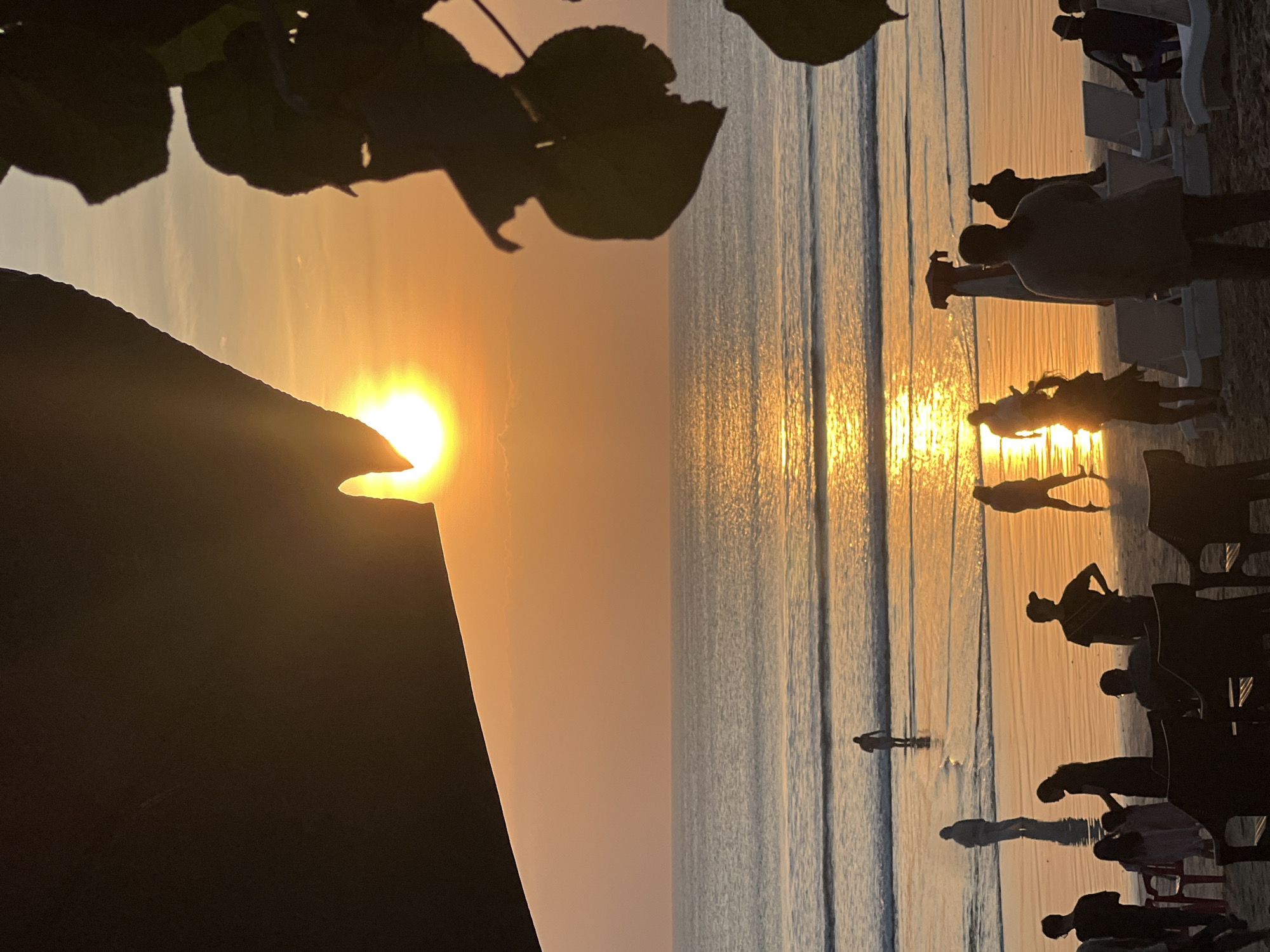 This is a photograph of the Kuta beach in Bali, Indonesia, taken during sunset. The sun is low in the sky, casting a warm orange glow across the scene and creating a shimmering reflection on the water. Silhouettes of people are visible on the beach and in the shallow water, some walking and others standing. The foreground is partially obscured by the dark silhouette of a large object, possibly a roof or an awning, and the leaves of a plant. Beach chairs are scattered on the sand. The ocean appears calm with gentle waves.