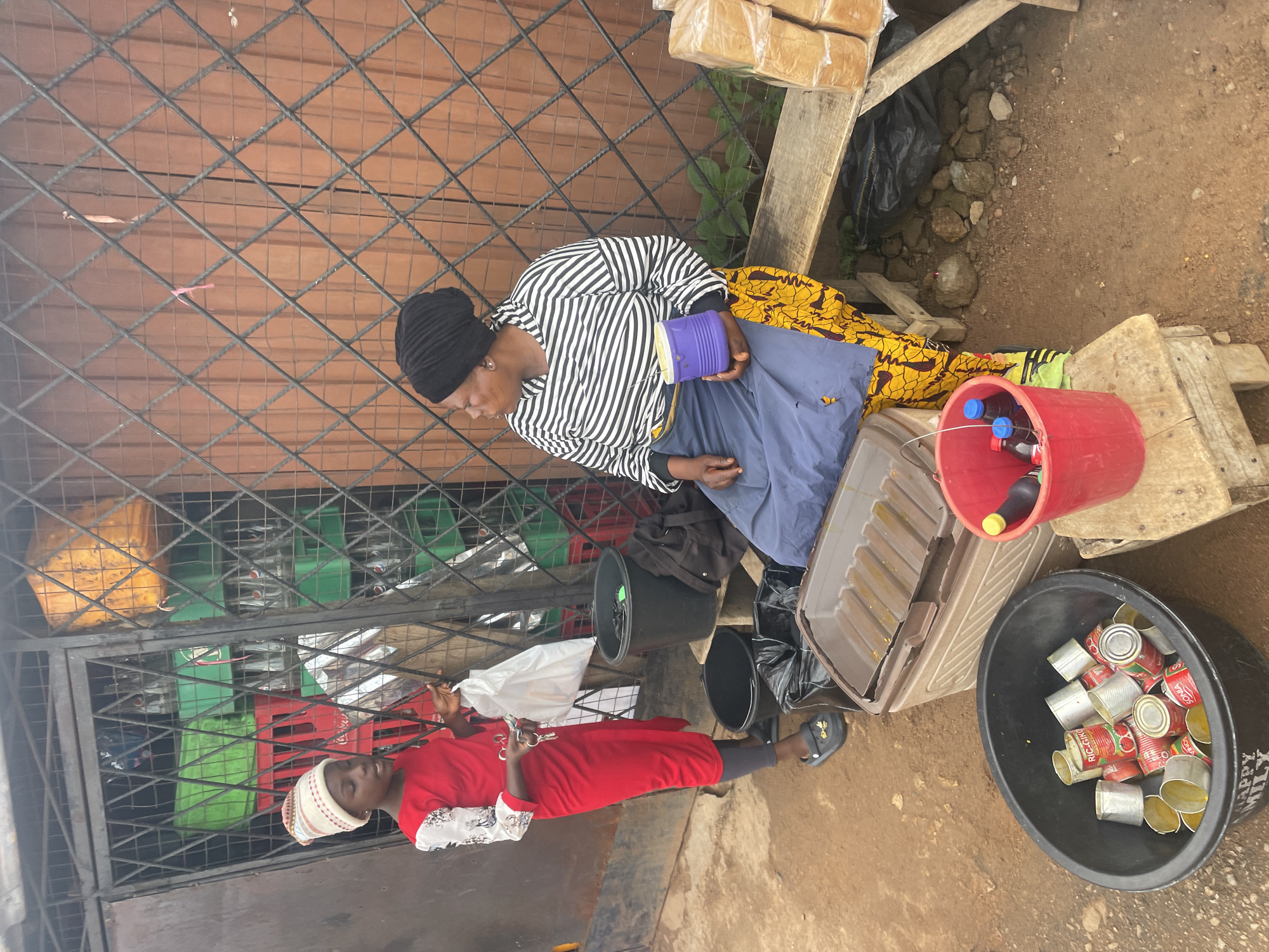 A young girl in a red dress and patterned beanie stands to the left of the frame, facing a woman in a black headscarf and striped shirt. The girl holds a white plastic bag and what appears to be keys in her right hand. The woman, seated on a wooden bench to the right, holds a purple container. Behind them, a metal grate covers a display of stacked crates and large yellow containers. The ground is a mixture of dirt and scattered rocks. To the foreground, a black bucket is filled with empty tin cans, and next to it, a large brown cooler. A red bucket containing bottles of liquid sits on a small wooden stool. Stacked loaves of bread are visible on a wooden shelf to the far right. The scene suggests an outdoor market or roadside stall in the city of Jos, Nigeria, characterized by its utilitarian setup and common street vending items.