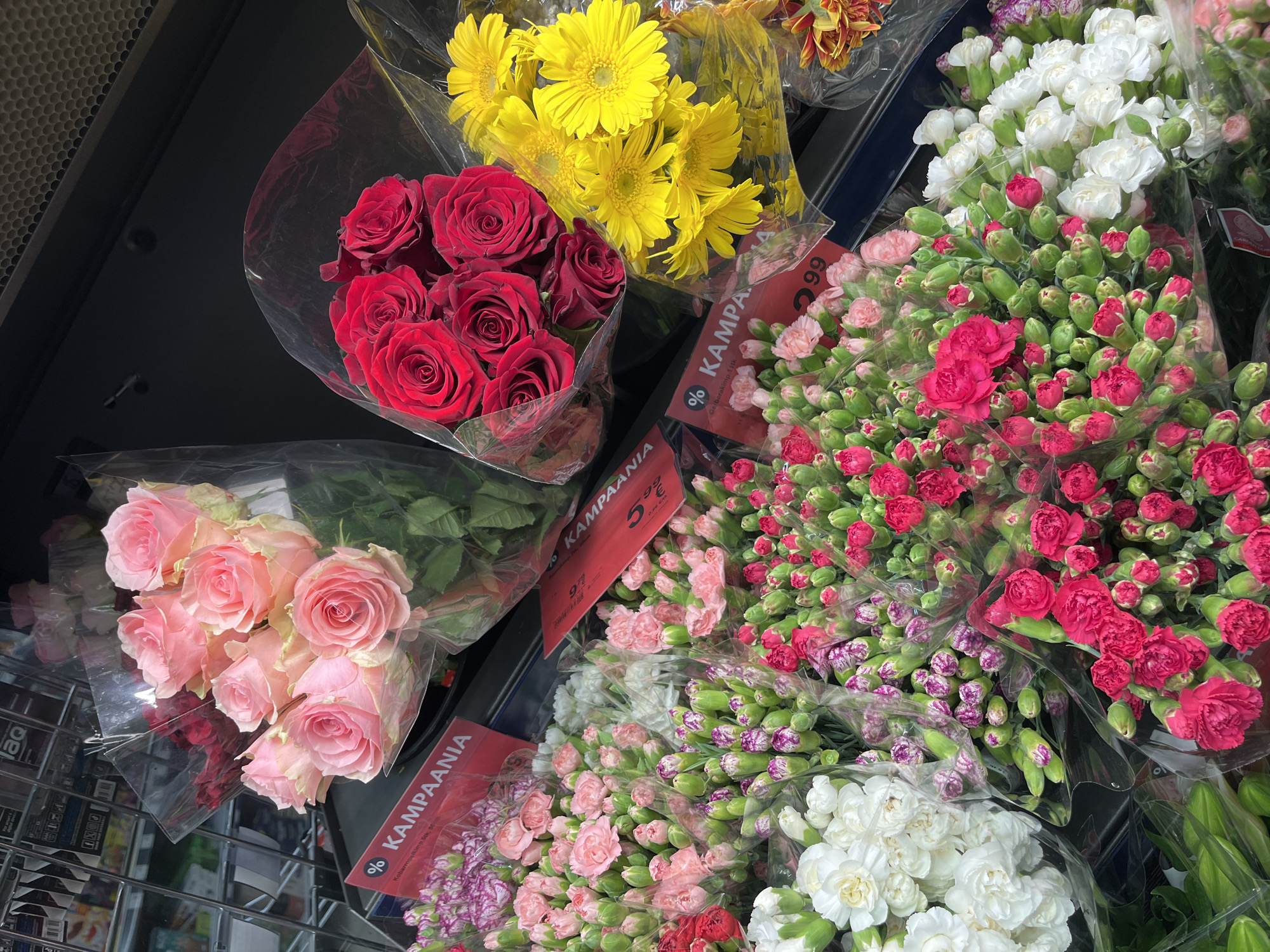 This is a close-up, eye-level, outdoor, daytime shot of a flower display at a market stall, possibly in the city of Kohtla-Järve, Estonia. The arrangement features several bouquets of flowers, predominantly roses and carnations, packaged in clear plastic.
In the foreground, there are abundant bunches of small pink and white carnations, with some vibrant red carnations also visible. The carnations are mostly in bud form, with a few blooms open. To the left and right of these, more mature blooms of white and pink carnations are present.
Further back, a bouquet of bright yellow gerbera daisies is prominently displayed, alongside a bouquet of deep red roses. Behind these, a bouquet of pale pink roses is visible, with some darker pink roses peeking out from behind.
Red signage with white text is interspersed among the flowers, some of which appear to be sale signs with "%" symbols and prices. The overall impression is one of a vibrant and colorful floral offering at a retail setting.