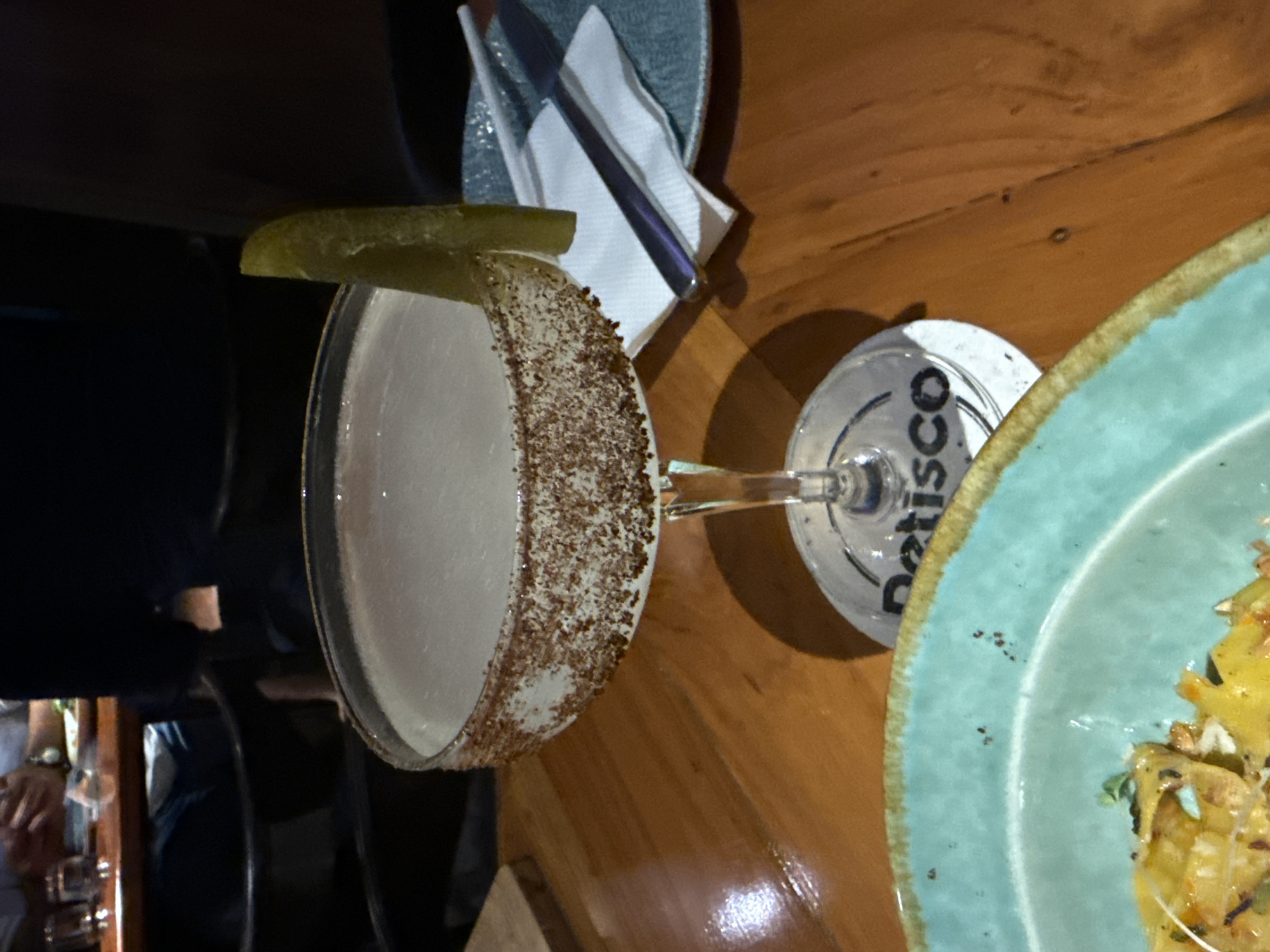 A cocktail glass with a spiced rim and a lime wedge sits on a coaster that says "Detisco", next to a plate of food. The items are on a wooden table in what appears to be a bar or restaurant setting. Other people are visible in the background. Based on the coaster, the location is likely Panaji, India.