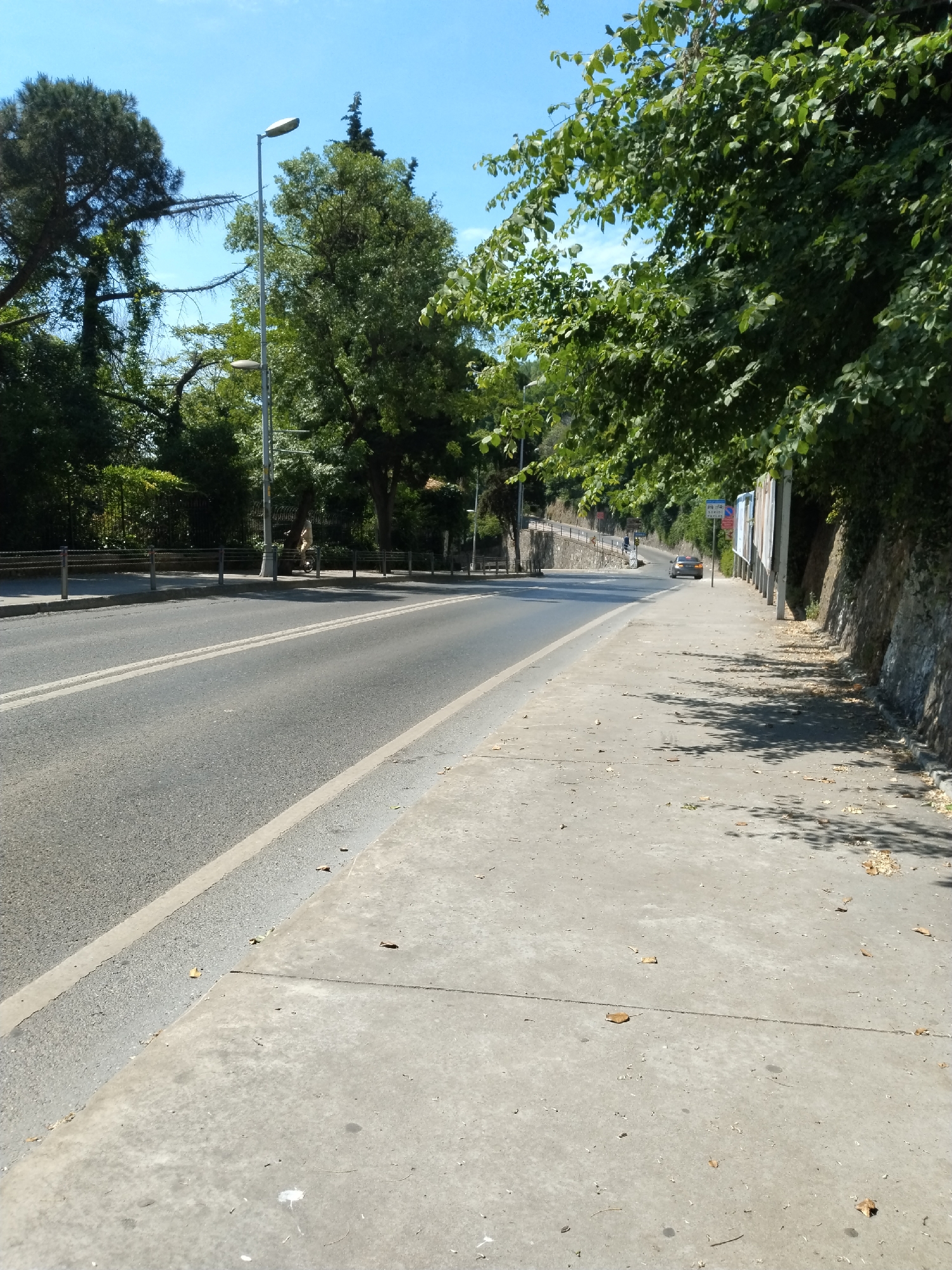 The image presents a sunlit roadside scene in Sarıyer, Turkey, under a clear blue sky. A two-lane asphalt road, marked with double and single white lines, recedes into the distance. A silver or light-colored car is visible driving away in the far distance.
To the right, a wide concrete sidewalk is partially covered by the dappled shadows of lush green trees overhead. Dry leaves are scattered across its surface. This side of the road is bordered by a low stone retaining wall and a row of large, faded billboards. A small blue road sign, bearing white pictograms (possibly a bus symbol) and text, which appears to include "TAKSI," stands near the billboards, likely indicating a bus stop or taxi stand.
On the left side of the road, dense green trees and bushes form a verdant backdrop. A tall street lamp with two lights is a prominent feature. Metal chain barriers line the edge of the road, behind which a person is seen riding a bicycle. Further down this side, two other figures are visible walking. The scene depicts a typical daytime setting for travel and pedestrian activity in a green, semi-urban environment, characterized by bright sunlight and strong shadows.