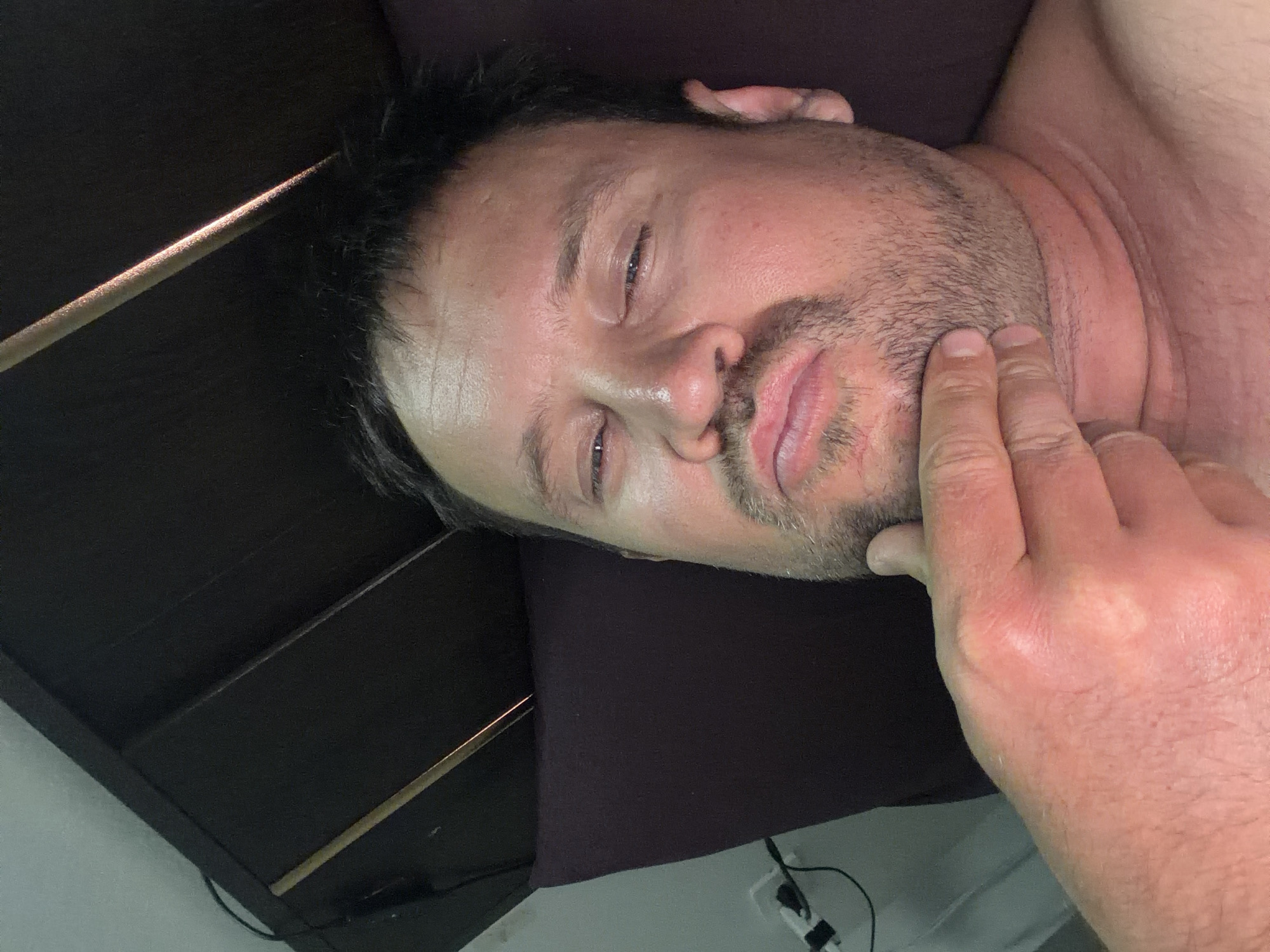 A close-up, low-angle selfie shows a Caucasian male lying down, propping his head with his left hand. The man has dark hair, a light complexion, and stubble on his chin. His eyes are partially closed, and his lips are pursed. He is wearing a dark shirt. The background consists of a dark-colored headboard, a pillow, and a wall with a light switch and electrical outlet. The scene appears to be a bedroom in Baan Mae Nam, Thailand.
