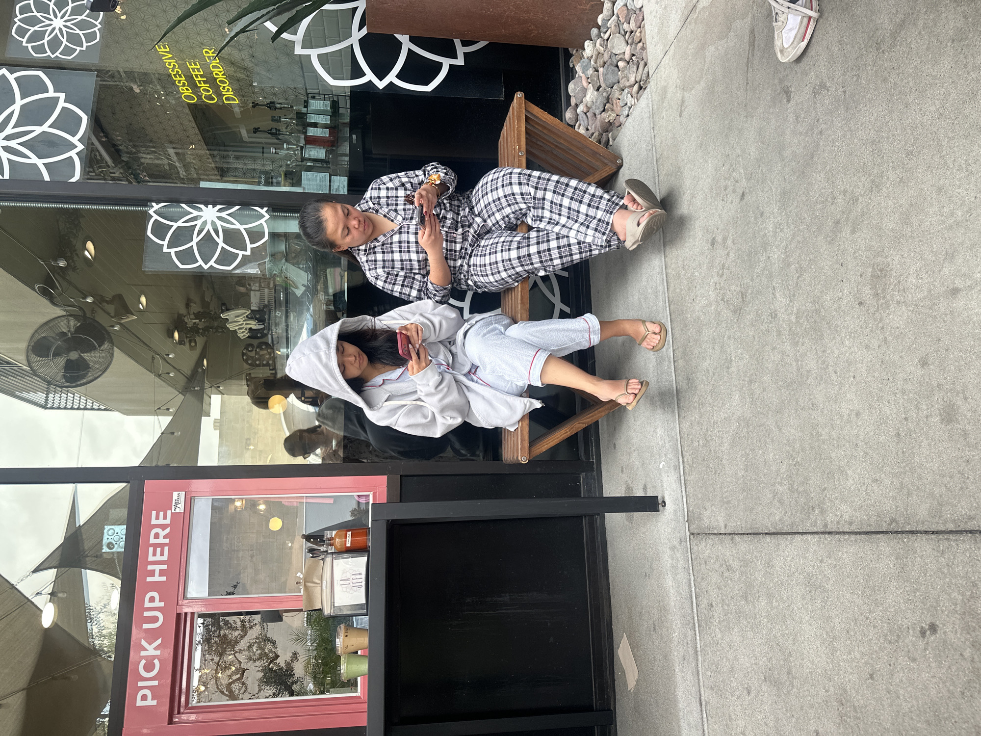 Outside a business in an unidentified city, two individuals wearing pajamas are seated on a bench, looking at their mobile phones. The location has a "PICK UP HERE" sign, and the window displays the phrase "OBSESSIVE COFFEE DISORDER". A third person's feet are visible, partially in the shot. The surrounding environment appears to be an urban street.