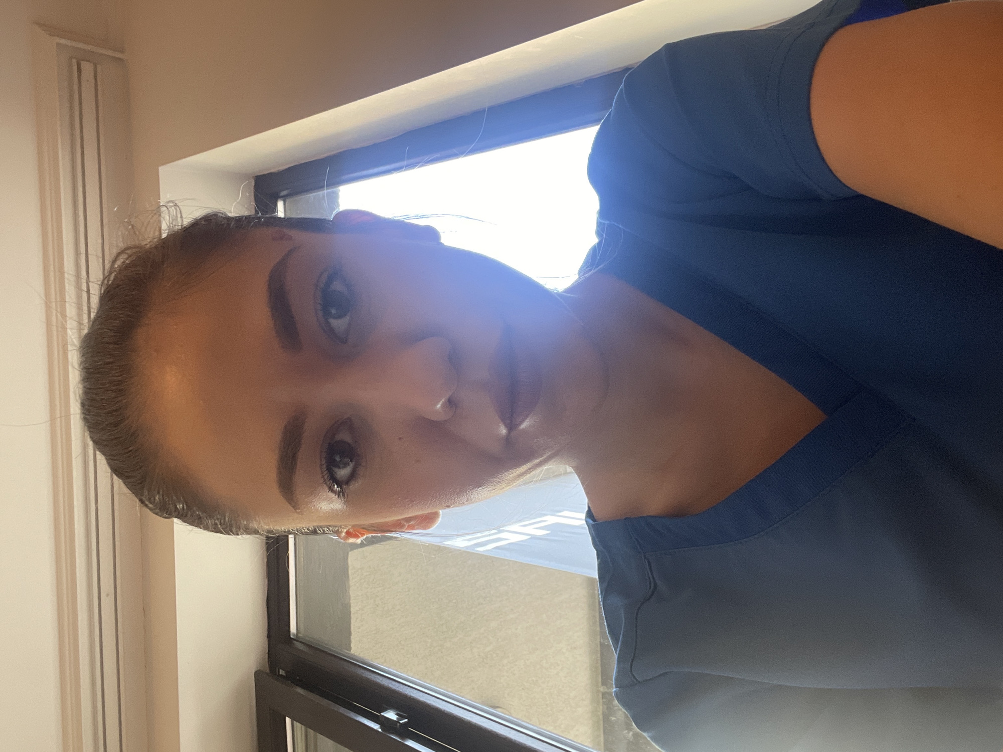 The image, rotated 90 degrees clockwise, depicts a close-up portrait of a woman, likely a selfie. She is an adult with dark hair pulled back and distinct eyebrows, appearing to look slightly upwards towards the camera. She is wearing a dark blue or teal scrub top, suggesting a professional medical or caregiving environment in Sofia, Bulgaria. Her expression is neutral.
The setting is indoors, with a prominent window behind her that floods the scene with intense daylight, causing significant backlighting and lens flare. The light indicates it is daytime with bright, clear weather. Through the window, the outdoor scene is overexposed, making it difficult to discern details. However, faint, blurry white text is partially visible on what appears to be an outdoor sign, but it is indistinct and cannot be clearly read (possibly "ES" or "FS" or "S"). To the viewer's left (her right), a light-colored wall with vertical grooves or molding is visible. The focus is primarily on her face and upper body, showcasing her attire and the strong, natural lighting.