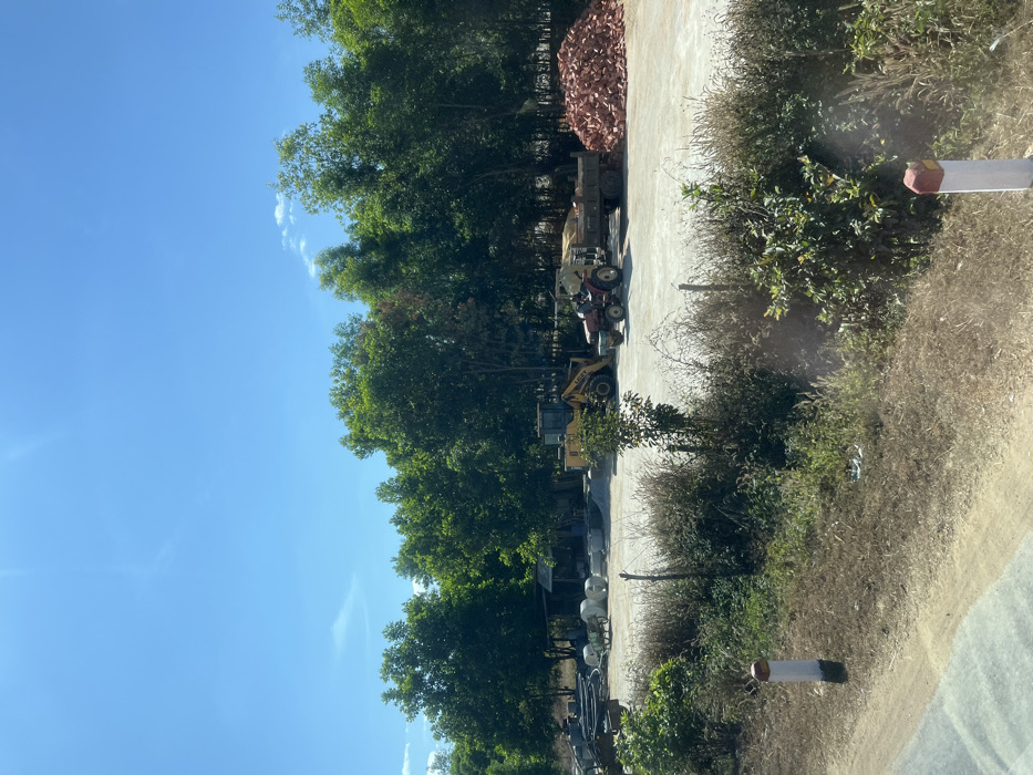 A vertical shot reveals a blue sky and tall green trees to the left. Various construction vehicles and equipment, including a bulldozer and tractor, are parked on a concrete surface in the center. To the right of the vehicles is dense, dry vegetation. A pile of reddish-brown material sits near the back, behind the vehicles. A white post with a red top appears in the lower right corner.
