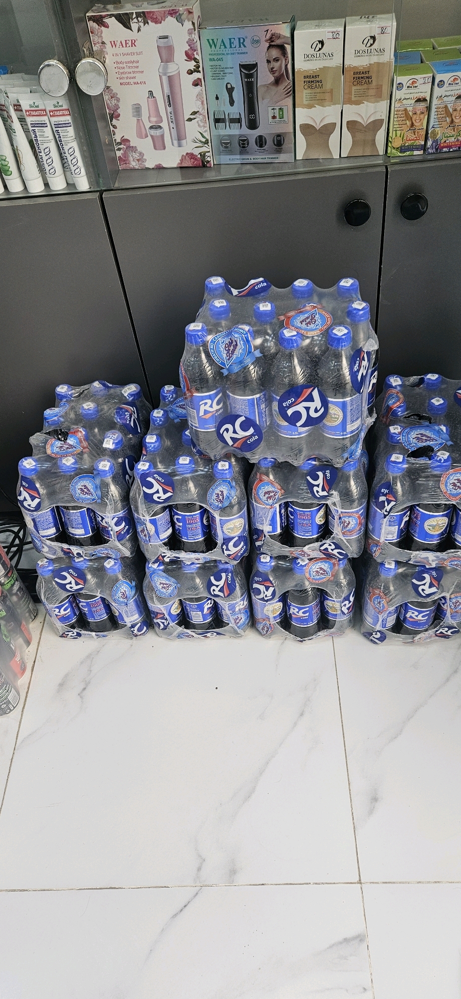 This image captures a retail display in Dushanbe, Tajikistan, featuring a variety of products. On a shelf, several boxes of personal care items are visible, including "WAER" branded electric shavers and "DOS LUNAS" breast firming cream. Below the shelf, multiple multipacks of "RC COLA" bottled drinks are stacked on the floor, shrink-wrapped for sale. The flooring is white with a marble-like pattern. The scene appears to be an indoor establishment, likely a shop or supermarket, showcasing consumer goods. No individuals are present in the image.