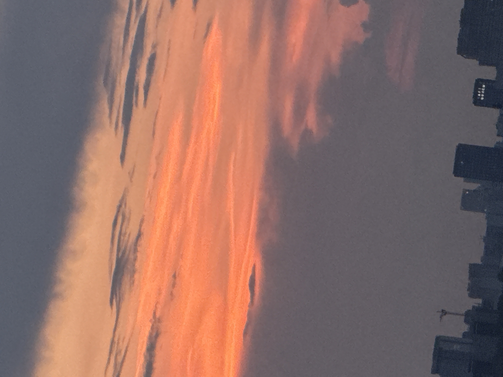 A cityscape is silhouetted against a dramatic sunset sky. The sky transitions from a deep, dusky blue at the top to vibrant oranges and pinks, with streaks of darker clouds cutting through the colorful expanse. In the lower portion of the image, the dark outlines of buildings rise from the horizon, suggesting a metropolitan area. One building features illuminated text. A crane is visible among the structures, indicating ongoing construction. The overall impression is one of an urban setting at dusk, with the natural beauty of the sunset contrasting with the man-made structures.