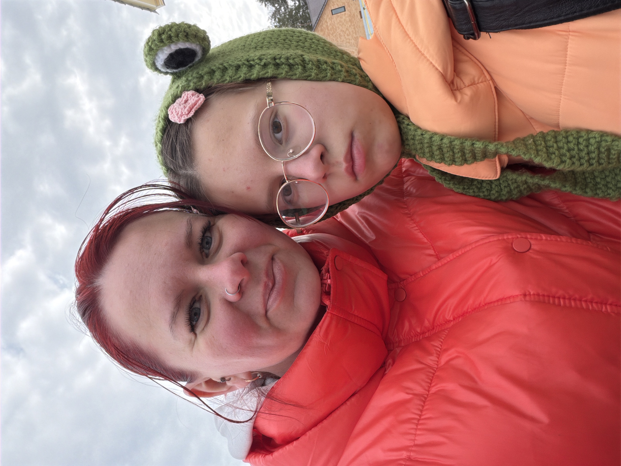 Two individuals are visible in the image. The person in the foreground has red hair and is wearing a red puffer jacket. The individual behind them is wearing an orange puffer jacket, green frog-themed hat, and round glasses. The setting appears to be outdoors under a cloudy sky, with a building visible in the background. While there is no direct evidence within the image itself confirming the location, the prompt specified that the setting is Chaikovsky, Russia.