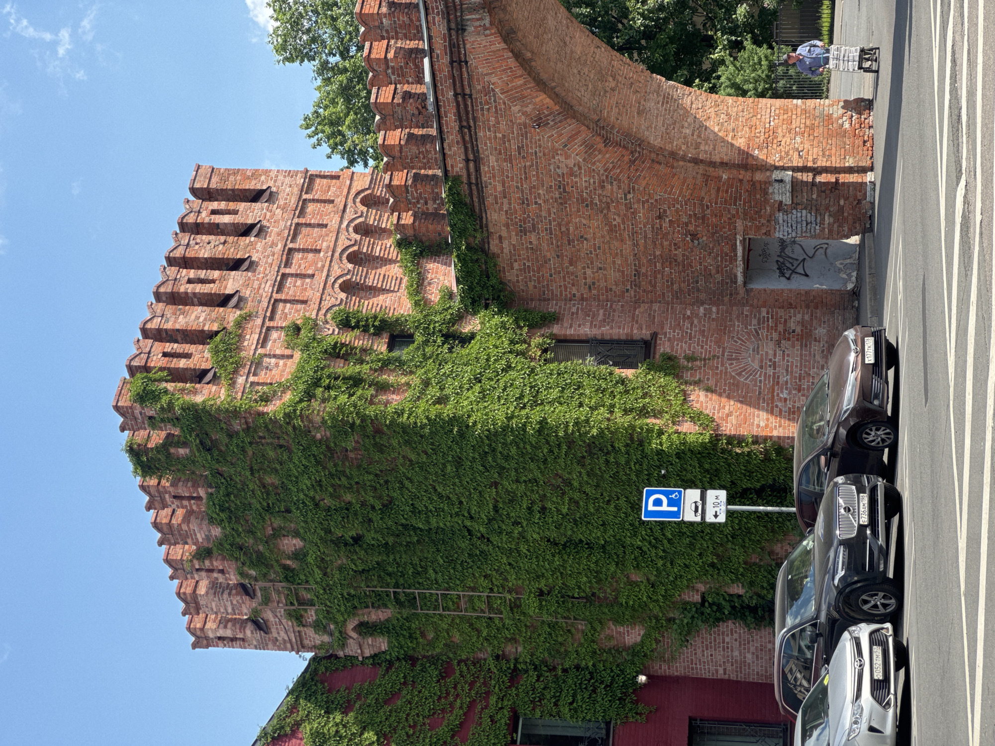 A daytime outdoor scene features a tall, historic-looking brick building heavily covered in green ivy under a clear blue sky. The building, possibly a tower or a section of a fortress wall, is constructed from reddish-brown bricks and exhibits architectural details such as a crenellated or stepped parapet at its top and arched, recessed sections. A metal ladder is visible ascending the ivy-covered wall.
To the right of the building, a paved street runs parallel, with several cars parked along its edge. The cars include a dark SUV (Volvo XC90) with a Russian license plate "E736KM 198" and a silver sedan with a partially visible plate "O052HP 98". Road markings, including white solid and dashed lines, are visible on the asphalt. In the upper right background, a person wearing a blue shirt is seen pushing a shopping cart on the sidewalk next to a curved brick wall and some trees.
A blue square parking sign is prominently displayed on the right side of the building, indicating "P" for parking, a disabled person symbol, and an arrow pointing left with "10 м" (10 meters) and a car symbol, suggesting a parking restriction or zone. Near the base of the brick wall, there's a recessed opening or doorway with some black and white graffiti, which is largely illegible. The bright sunlight suggests a clear, sunny day, with distinct shadows cast by the building's features. The architecture and license plate codes suggest a location in Russia, specifically Saint Petersburg due to the "198" and "98" region codes.