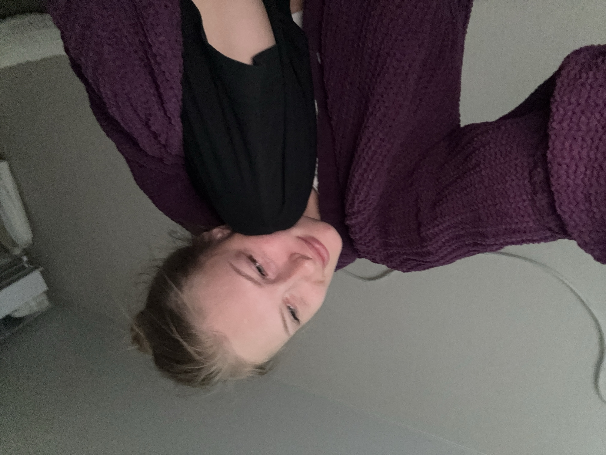 A young woman with blonde hair pulled back in a bun is seen in a close-up, head-and-shoulders shot. She is wearing a thick, deep purple knitted sweater or robe. Her head is tilted to her left, and she is looking upwards and to her right with a soft, contented expression. She is also wearing a black garment that is partially visible under the purple sweater. The background is a plain, light gray wall. To the right side of the frame, a portion of a shelf with some white items is visible, suggesting the setting might be a bedroom or a closet. There is a thin, light-colored cable or cord running across the upper left portion of the image. No specific landmarks or elements are present to indicate the city of Perm, Russia, or any other specific urban location.