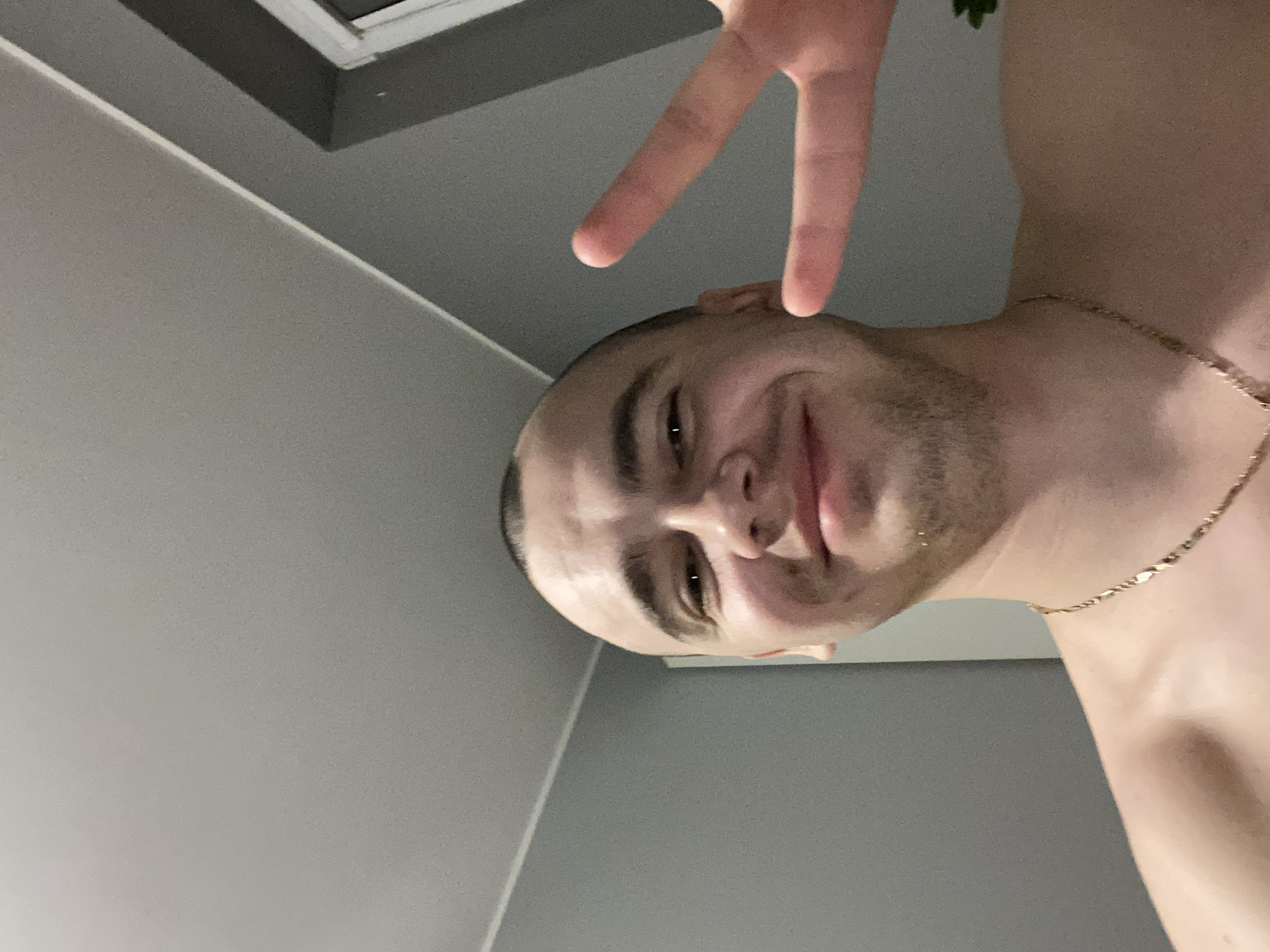 The image is a selfie of a young man with a shaved head, smiling and holding up two fingers in a peace sign. He is wearing a gold chain around his neck. The background appears to be an indoor setting with grey walls and a white ceiling. There is a hint of a window frame in the upper right corner. The lighting suggests it might be evening or a dimly lit room. The subject's pose and expression are casual and friendly. There is no discernible information about the specific location within Łódź, Poland.