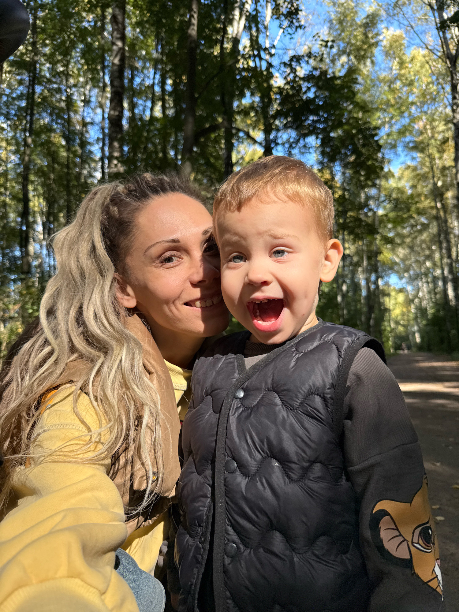 A close-up, vertical, selfie-style shot features a woman and a young child posing in a sun-dappled forest in Rusanovo, Russia. The woman, with blonde hair pulled into a high ponytail, smiles broadly, her head tilted towards the child. She wears a yellow hoodie and a brown scarf. To her right, the child, with light brown hair, looks directly at the camera with an open mouth and wide eyes, appearing excited. The child is wearing a dark, quilted vest over a dark gray long-sleeved shirt with a lion graphic on the sleeve. Behind them, the forest is illuminated by sunlight filtering through the leaves of tall trees, creating a blurred background of green and brown. The ground in the background appears to be a dirt path.
