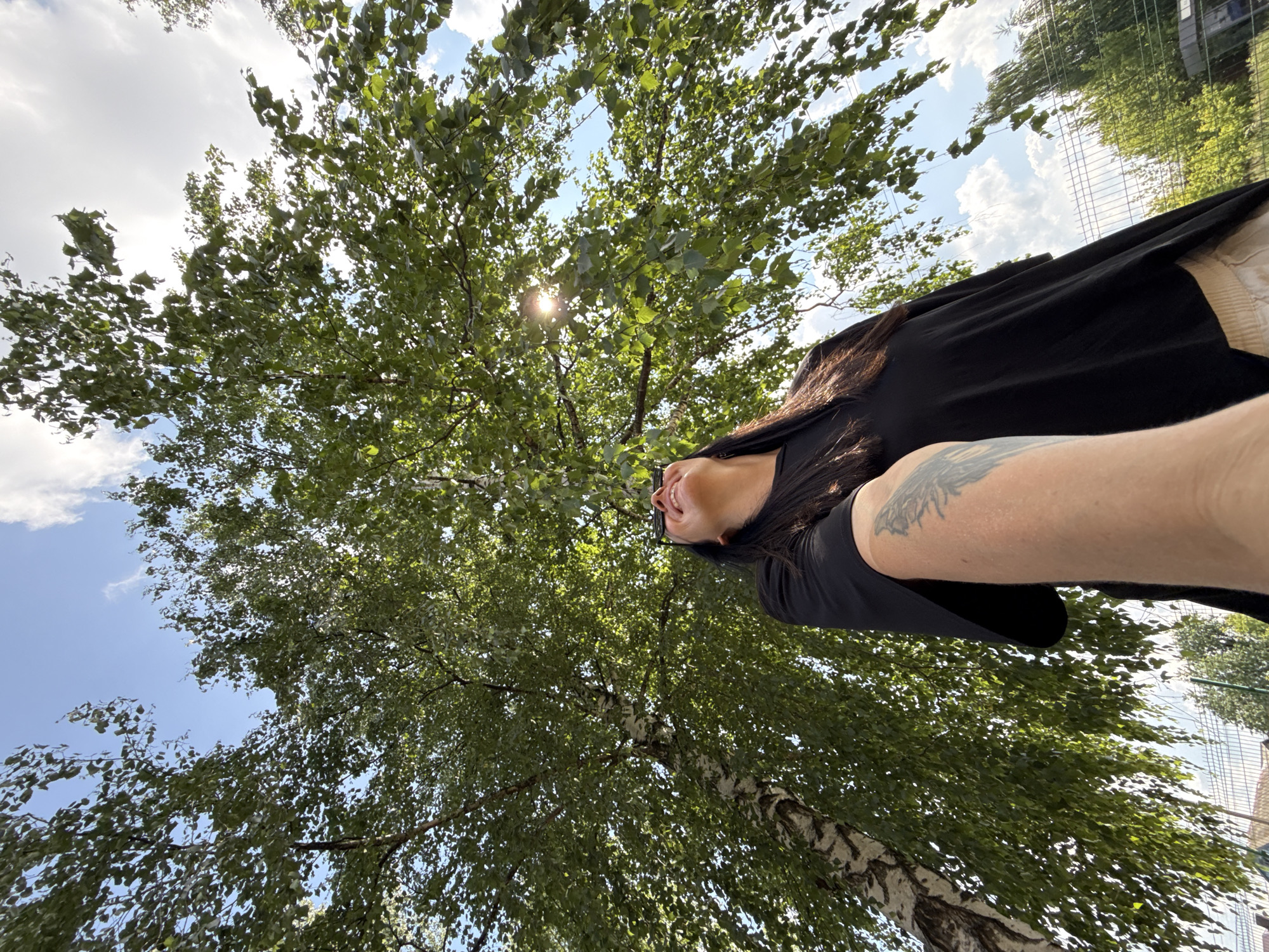 A woman is looking up at a large tree with green leaves, her face illuminated by the sun. She is wearing a black t-shirt and light-colored shorts. Her arm is extended, holding a camera or phone, suggesting she is taking a selfie. The background shows a bright blue sky with scattered clouds and a metal fence. The scene appears to be outdoors, possibly in a park or residential area, given the presence of the fence and a glimpse of buildings in the distance. The image is taken from a low angle, emphasizing the size of the tree and the sky. The location is specified as Chaikovsky, Russia.