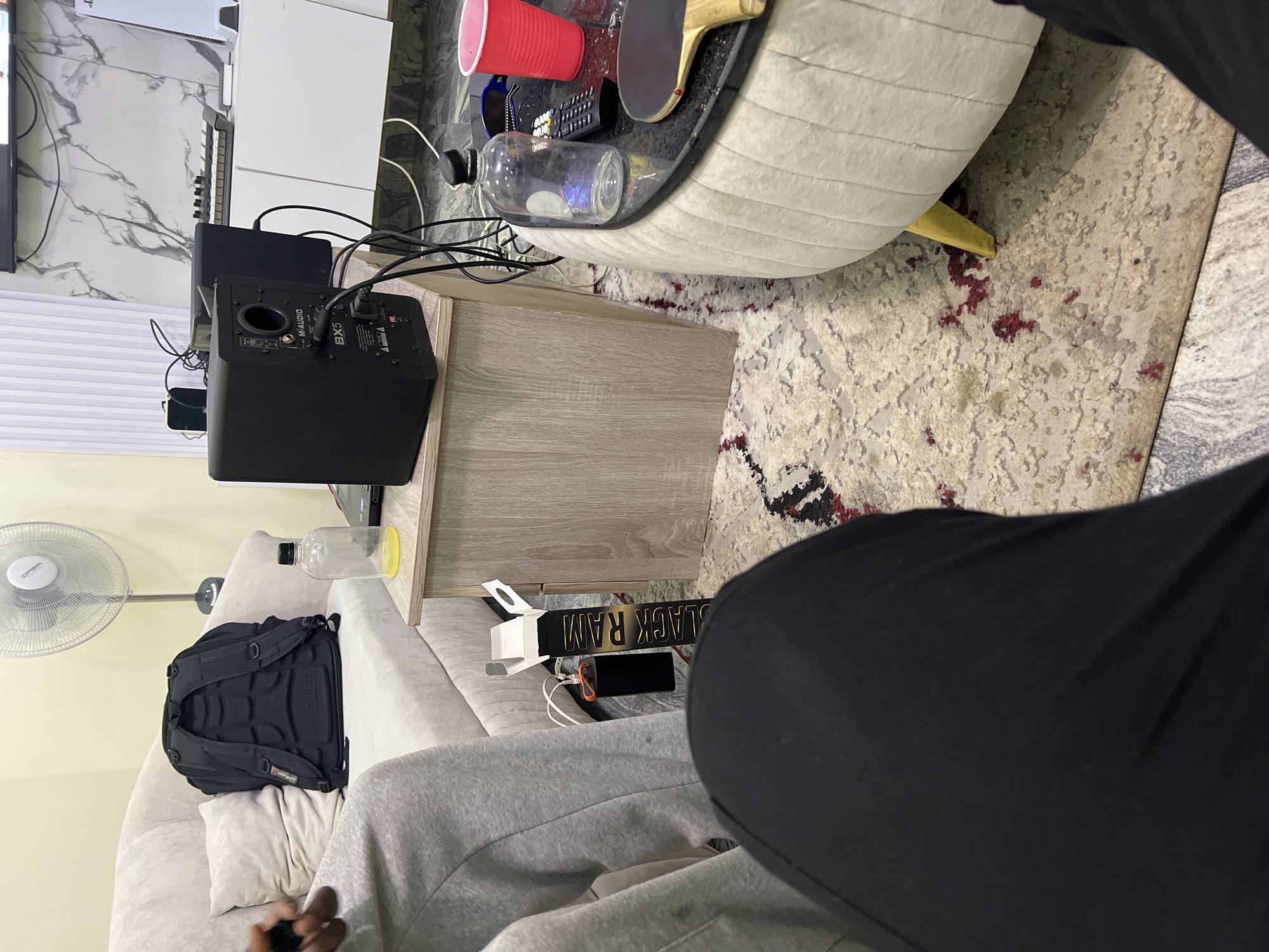 The image, taken in Gwarinpa, Nigeria, depicts an indoor living space from a first-person perspective, looking down and across a room. In the foreground, the legs of a person wearing grey sweatpants and black trousers are visible, with hands holding a dark object, possibly a phone or remote.
The mid-ground features a casual living area. To the right, a light-colored couch holds a black backpack and a white pillow. Next to the couch on the floor, a power bank with white cables and a book with "BLACK RAM" on its spine are visible. A white standing fan is positioned to the left of the couch. To the left of the scene, a black M-Audio BX5 studio monitor speaker rests on a light wooden side table, accompanied by another speaker and a tangle of wires.
In the center of the mid-ground is a light-colored, upholstered coffee table with a dark top, holding a red solo cup, a clear plastic bottle with a black cap, a remote control, sunglasses, and a ping-pong paddle.
The background reveals a white wall with a grey marble pattern. Against this wall, a television screen is partially visible on the far left, above a white PlayStation 5 console and a musical keyboard/MIDI controller. The floor is covered by a light-colored patterned rug featuring red accents, with a section of grey tiled floor visible on the far right. The room appears lived-in, suggesting a relaxed domestic setting. The visible text includes "M-AUDIO" and "BX5" on the speaker, "OPERA" on the fan, "WPPWER" (or similar) on the backpack, and "BLACK RAM" on the book. The time of day appears to be daytime, with general indoor lighting.