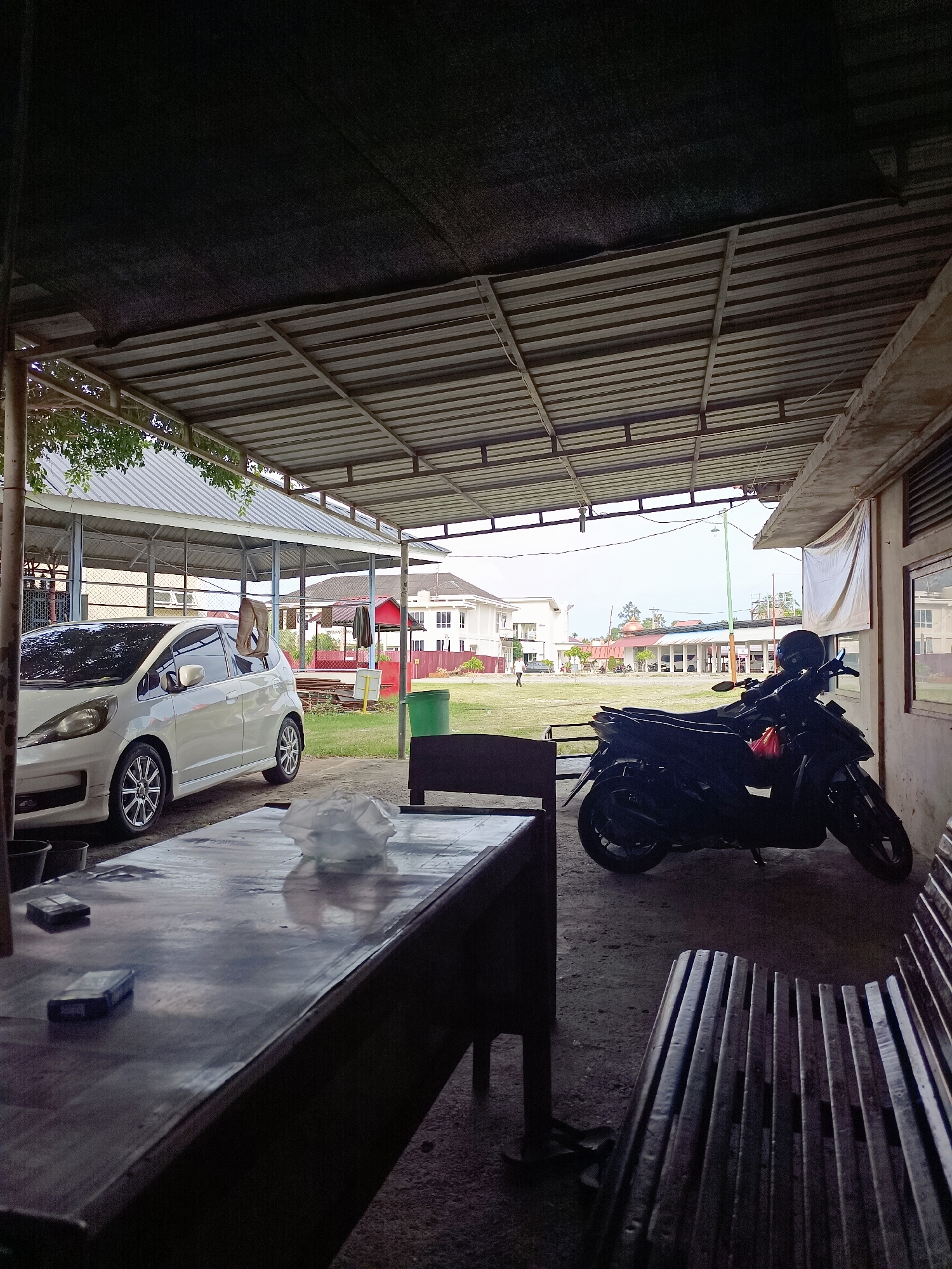 In Mesjid Reuntoh, Indonesia, this image captures a covered outdoor area, possibly a waiting lounge or parking space, with a rough concrete floor. A white hatchback car is parked to the left, its front passenger side visible. To the right, a dark-colored scooter with a black helmet on its handlebars is parked. In the foreground, a long, dark wooden table with a shiny surface is positioned, holding a crumpled white plastic bag and two rectangular dark objects, possibly cigarette packs. To the right of the table, a dark wooden bench with a slatted seat is visible. Beyond the covered area, a grassy field stretches out, with several low-rise buildings in the background, including structures with red roofs and white walls. The sky is overcast.