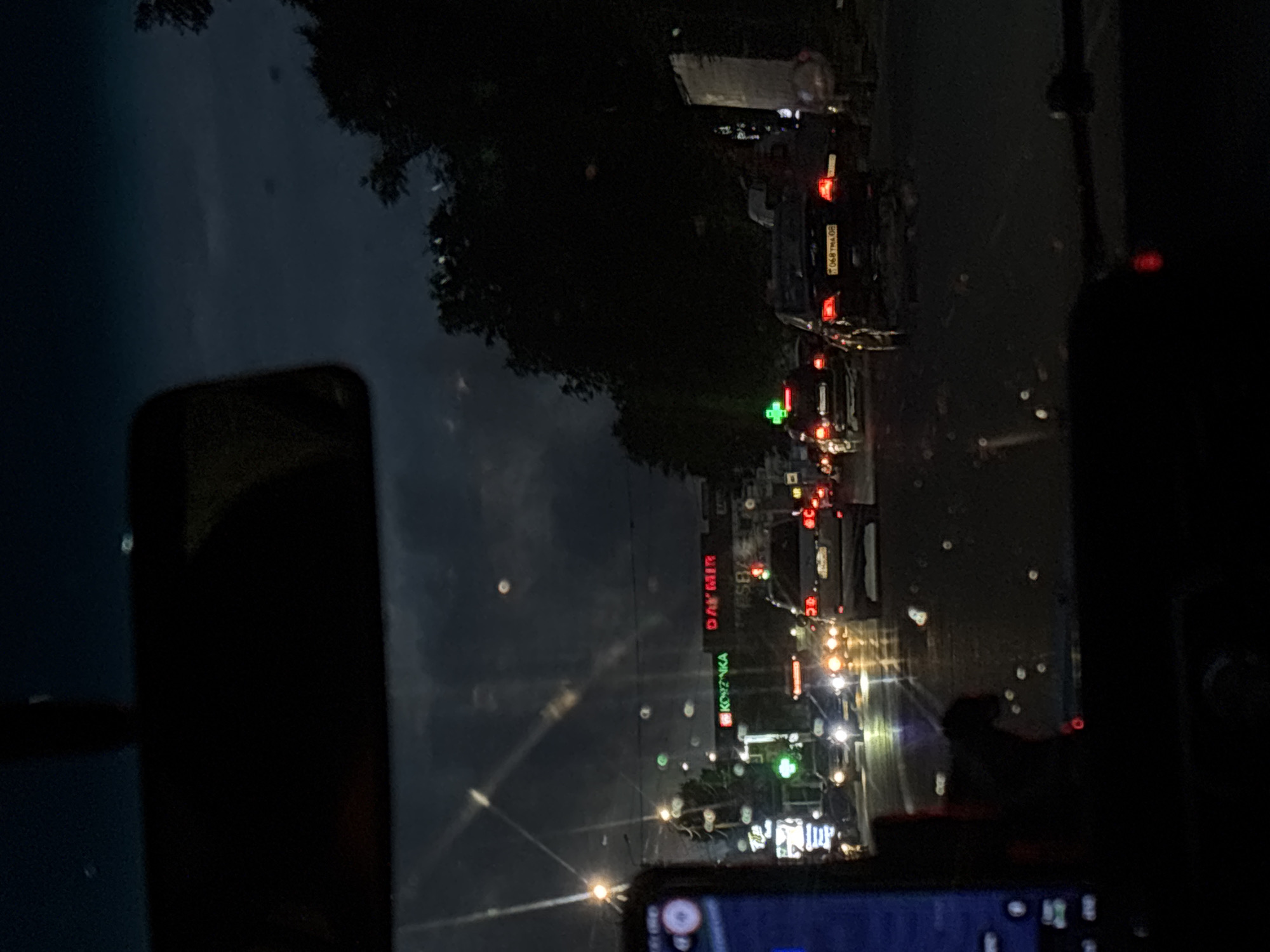 The image is a night-time photograph taken from inside a vehicle, looking out through the windshield. The view shows a street scene with traffic, buildings, and trees under a dark, cloudy sky. In the foreground, a part of the vehicle's interior is visible, including what appears to be a phone mounted on the dashboard on the left.
The street is wet, indicating recent rain or a wet road surface, with reflections of lights visible. Several cars are on the road, some with their headlights and taillights on. Traffic lights are visible, one showing green and others obscured.
On the right side of the street, there are buildings with illuminated signs. One sign reads "KÖZUKA" in green lettering, and another sign to its right says "DAYMIR" in red lettering. There are also other illuminated signs and windows on the buildings.
On the left side of the street, there are more buildings and streetlights. A green cross symbol, often associated with pharmacies, is visible on a building.
The overall impression is a typical city street at night, possibly during or after a rain shower, with moderate traffic. The scene appears to be set in an urban environment. The prompt mentions Taraz city administration, Kazakhstan, but there are no specific visual cues in the image to confirm this location.