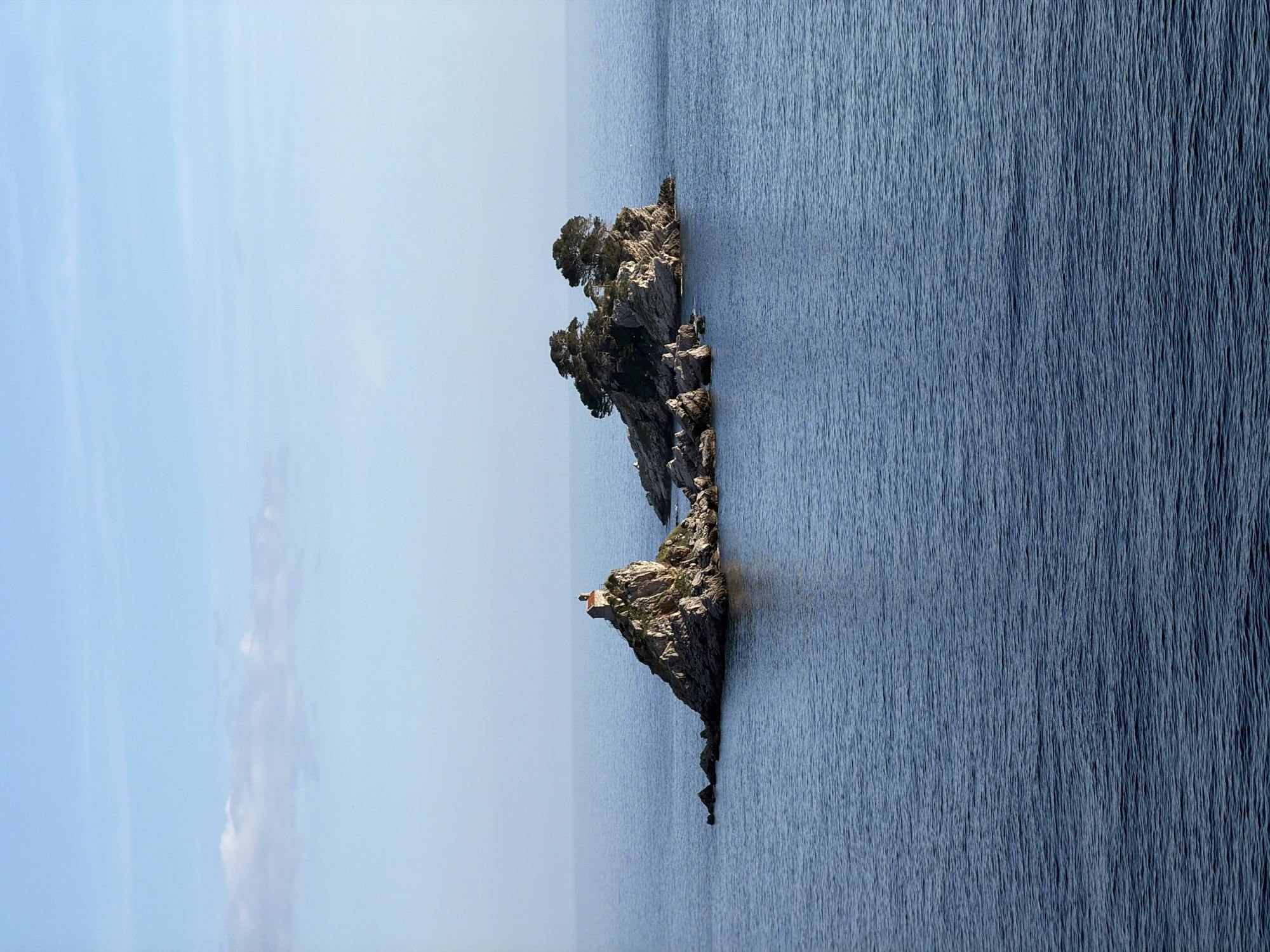 The image shows a rocky islet in the Adriatic Sea off the coast of Petrovac na Moru, Montenegro. The islet features craggy rocks with sparse vegetation, including trees and shrubbery. On the smaller, lower part of the islet sits a small structure, possibly a chapel, with a red roof. The water surrounding the islet is deep blue with subtle wave patterns, and the sky is a pale blue with a few wispy clouds. The photograph is taken from a slight angle, making the island appear vertically oriented in the frame.