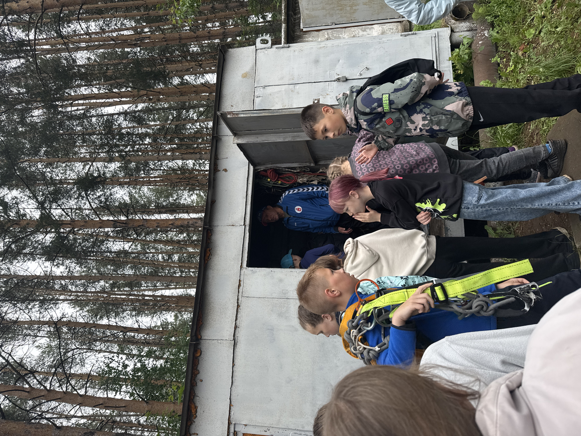 This image captures an outdoor scene featuring a group of children and teenagers gathered around a utilitarian, grey metal structure, possibly a storage shed, against a backdrop of dense trees. The image is oriented vertically, showing the subjects and the structure as if viewed from above, with the trees appearing horizontally in the background.
The subjects, mostly young people ranging from pre-teens to teenagers, are dressed in casual attire suitable for outdoor activities. Several individuals are visible in various states of readiness. A boy in a camouflage-patterned jacket, bearing the word "FEARLESS" on its sleeve, stands prominently, looking towards the open doorway of the structure. Another young person, closer to the foreground, is wearing a bright yellow and green safety harness with carabiners and chains attached, implying participation in a climbing or rope-course activity; part of the word "GRIP" is visible on the harness.
Through the open door of the metal structure, ropes, cables, and other equipment are visible inside. Several people are seen peering into or standing near the entrance, including an individual in a blue jacket with a "HUMMEL" logo, suggesting they might be receiving equipment or instructions.
The setting appears to be a wooded area or forest, with numerous tall, slender trees (possibly pines) forming a dense natural backdrop. The ground immediately around the structure shows patches of grass and earth. The lighting suggests it is daytime, likely overcast given the lack of strong shadows. The overall impression is one of an outdoor adventure or training activity, with participants preparing for an event. The location is Chaikovsky, Russia.