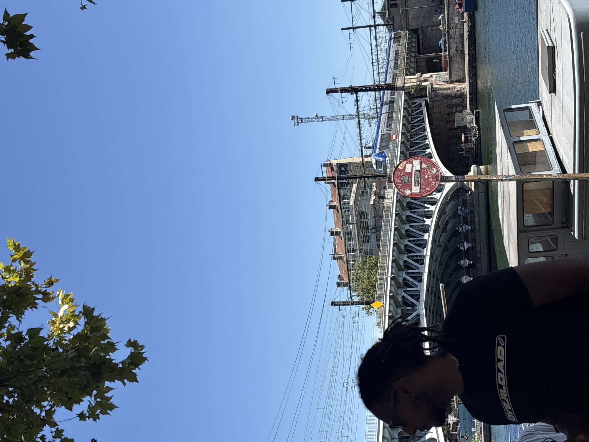The image captures a bright, sunny day with a clear blue sky over the Saône River in Lyon, France. In the foreground, a person with dreadlocks, wearing a black t-shirt with the word "OVERLOAD" visible, is looking to the left. Partially obscuring them is a part of a boat or barge.
Behind the person, a large, arched bridge spans the river, carrying a blue and white train. The bridge is an impressive structure of white metal with intricate arch supports. Along the riverbank, buildings with red-tiled roofs and several trees are visible. A tall communication tower with lights stands behind the bridge.
A red circular sign with white symbols, indicating a prohibition, is mounted on a pole in the middle ground, positioned in front of the bridge. Power lines and utility poles crisscross the sky above the train and bridge, suggesting an active urban environment. The water of the Saône River reflects the blue sky and the surrounding structures.