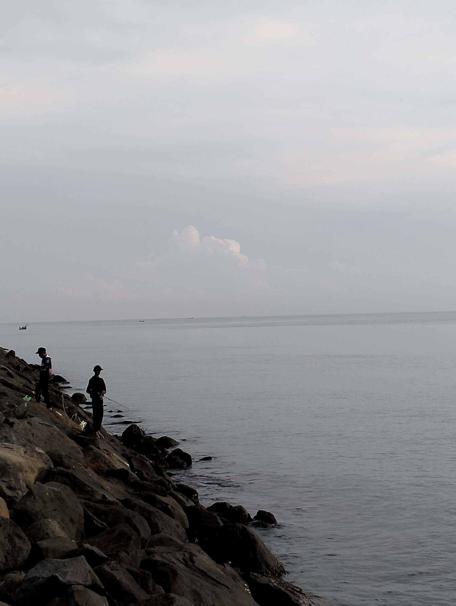 Two individuals are positioned on a rocky shoreline, likely engaged in fishing. The shoreline is constructed of large, dark boulders. The ocean stretches out behind them, meeting a gray, overcast sky. In the distance, faint outlines of boats can be seen. The location is likely near Lhokseumawe, Indonesia.