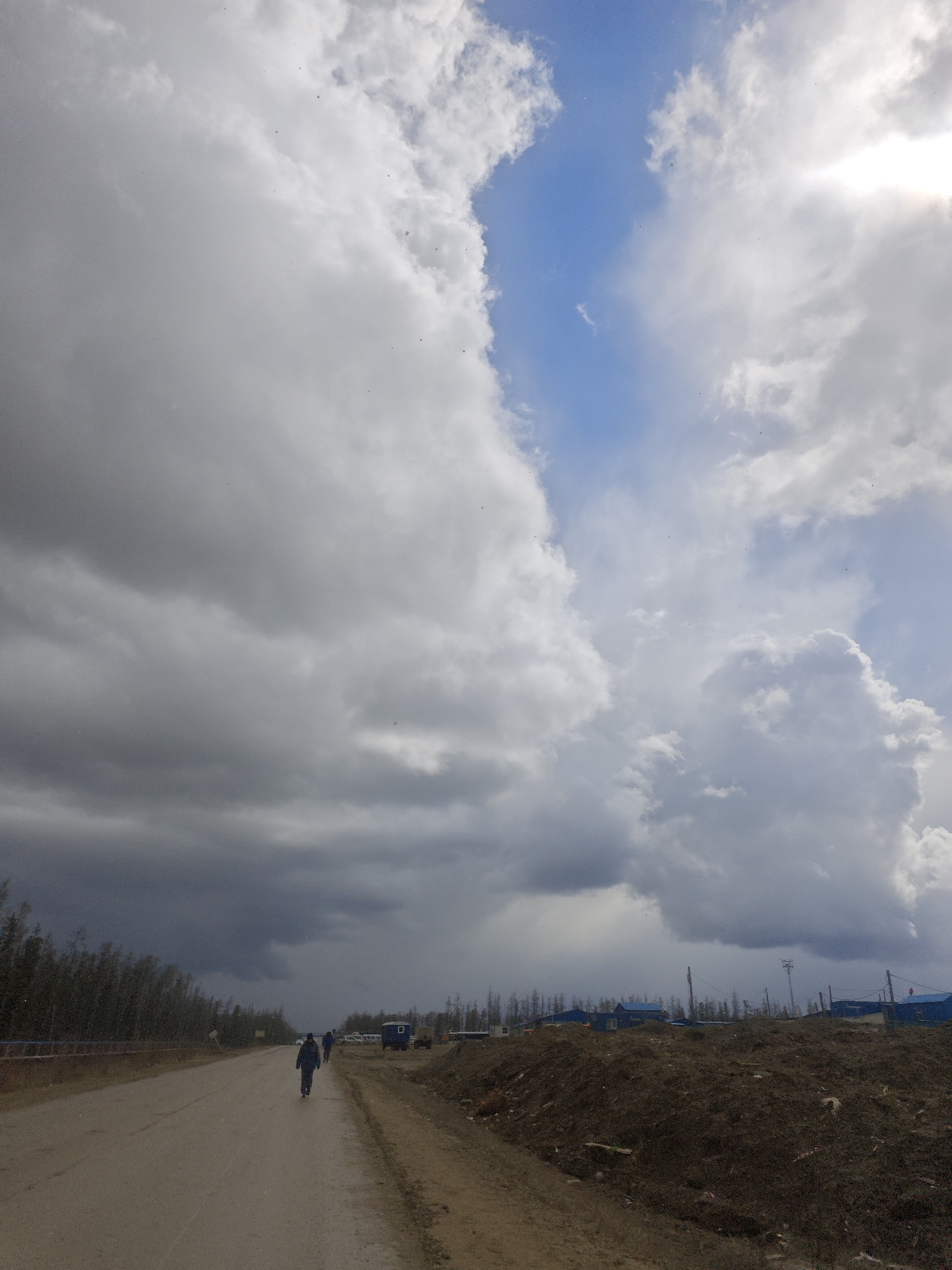 A wide-angle, ground-level shot shows a road in what appears to be a rural or semi-industrial area in Unknown, Russia. Two people are walking away from the viewer on the left side of the road. There are several vehicles, including a blue trailer-like structure, parked further down the road on the right side. To the left of the road, there is a line of thin trees. To the right of the road, there is a dirt bank. The sky is predominantly cloudy with a small patch of blue visible in the upper center. The sun is partially visible through the clouds in the upper right.