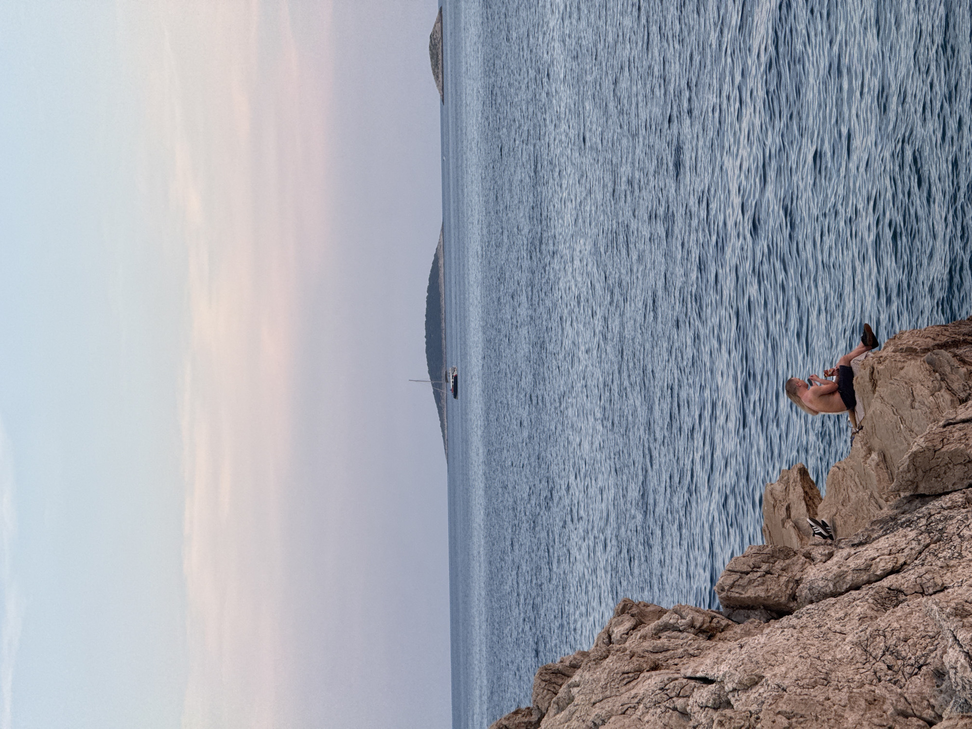 In Primošten, Croatia, a person sits on the edge of a rocky cliff overlooking the Adriatic Sea. The individual, with long blonde hair and bare torso, is wearing dark shorts and appears to be holding a drink. Their legs are crossed, and they are facing towards the right side of the frame, towards the water. To their left, on the rock, are a pair of black shoes. The sea is a deep blue with gentle ripples, and in the distance, a sailboat is visible on the horizon. Two small, tree-covered islands are also present on the horizon. The sky above is a soft gradient of pale blue and pink, suggesting either dawn or dusk. The rocky cliff face, occupying the left side of the image, is textured and reddish-brown in color.