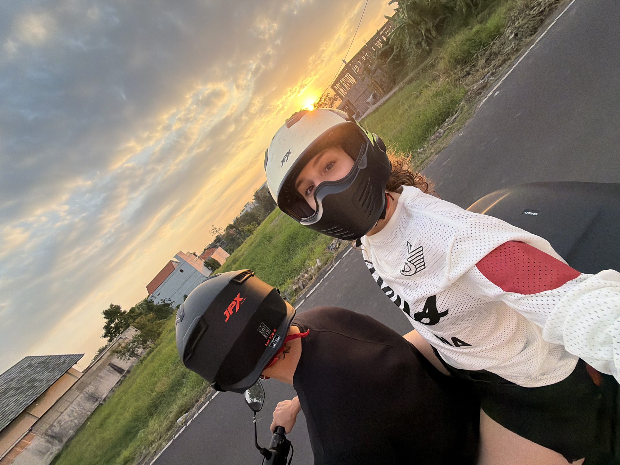 Two individuals are on a motorcycle, with the rider in the foreground taking a selfie, as they ride along a road in North Kuta, Indonesia, during sunset. The rider in the foreground is wearing a white mesh long-sleeved shirt with black and red accents and a cream-colored full-face helmet with a black visor and chin guard, featuring a "JPX" logo. Their eyes are visible through the helmet visor, and they have curly brown hair. The person behind them, likely the driver, is wearing a black t-shirt and a black full-face helmet with a red "JPX" logo. They are holding the motorcycle's handlebars. The background shows a rural or suburban landscape with green fields, a building with a tiled roof, and more structures in the distance. The sky is filled with dramatic, orange and grey clouds illuminated by the setting sun, casting a warm glow over the scene. The road surface is asphalt.